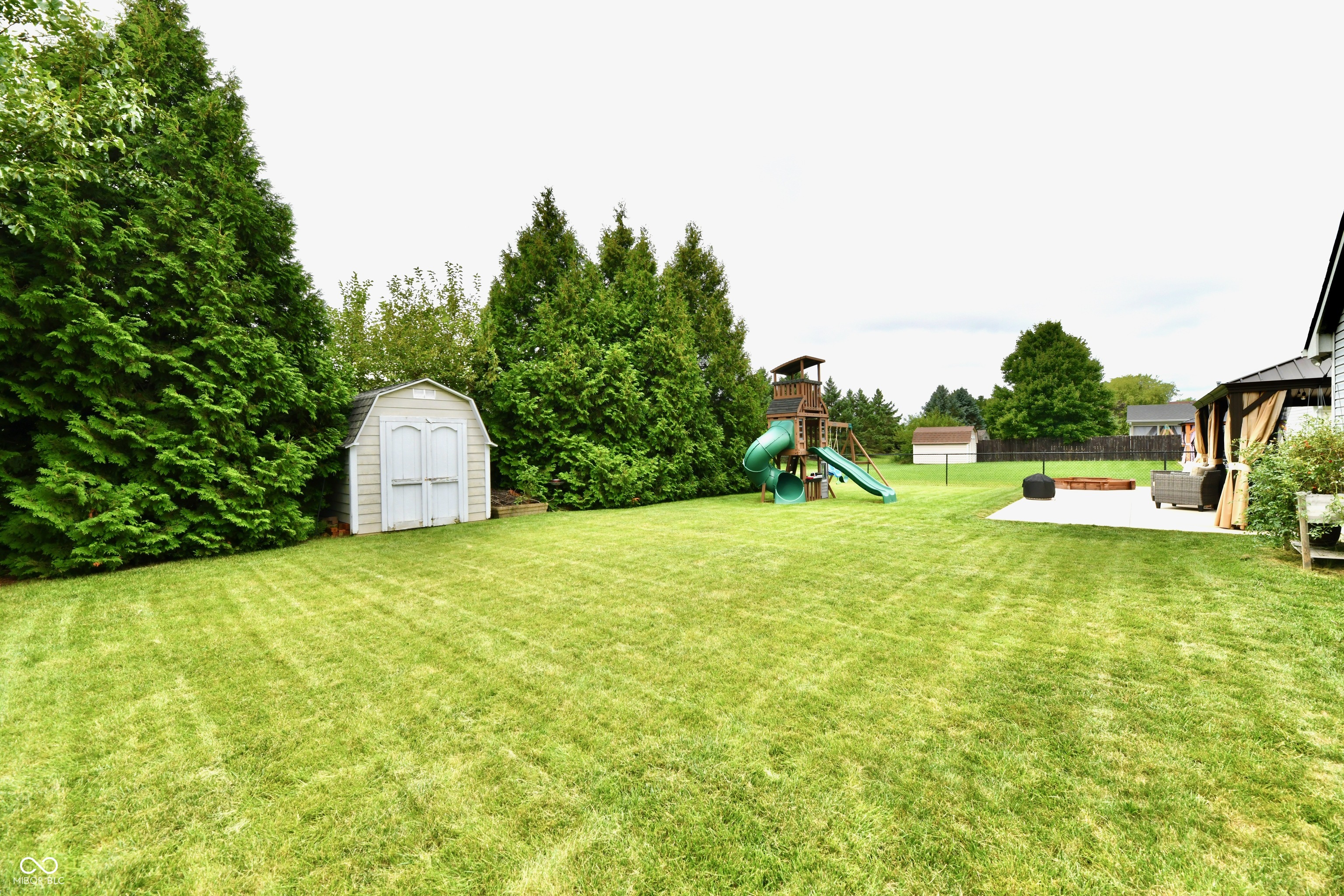 Backyard with a wide lawn, white shed, playset, and covered patio seating area.