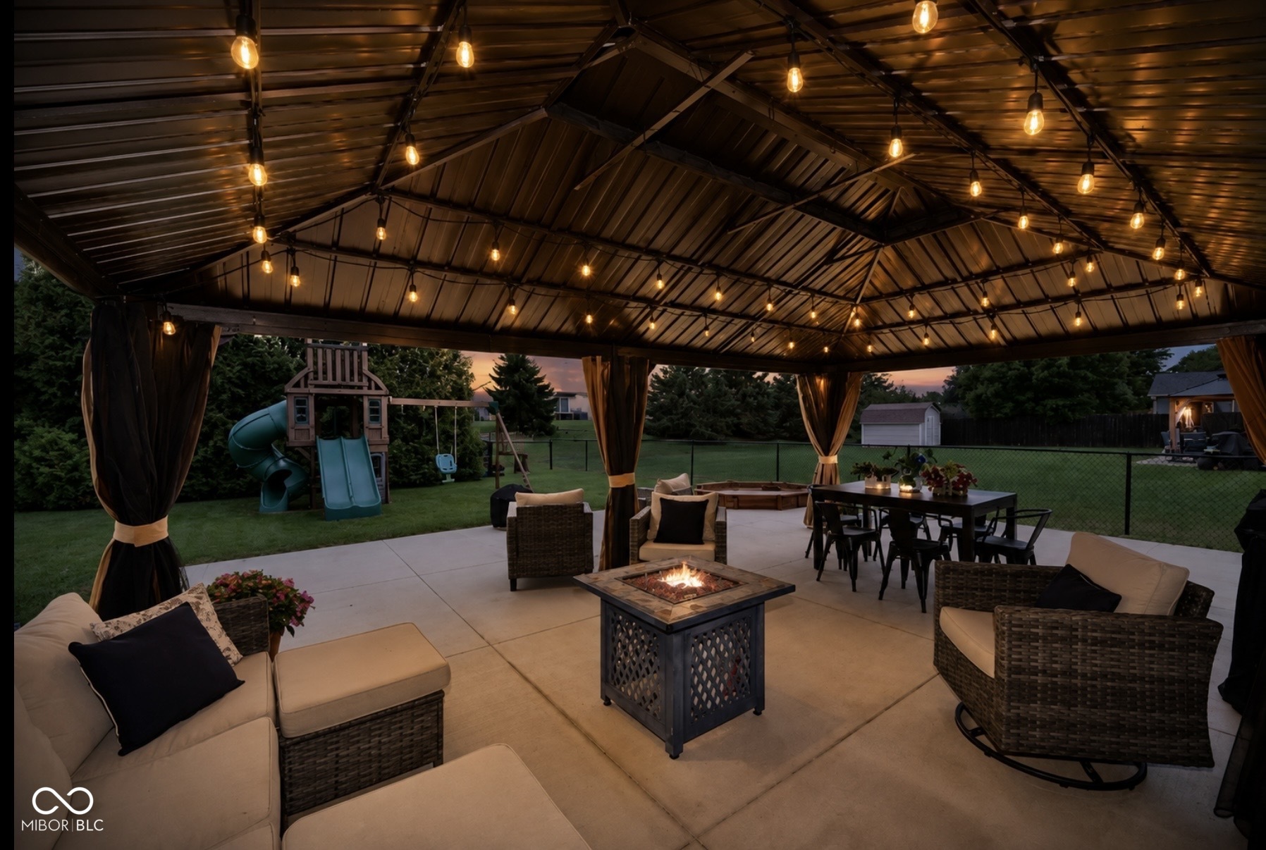 Covered backyard patio with string lights, wicker seating, dining table, and fire table at dusk.