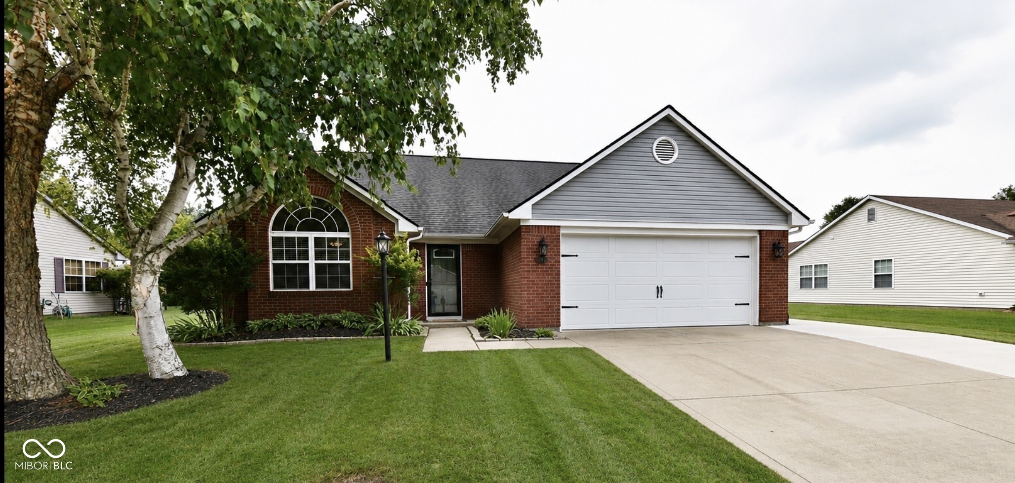 Brick home with gray gable siding, a white two-car garage, and mature trees across the front lawn.