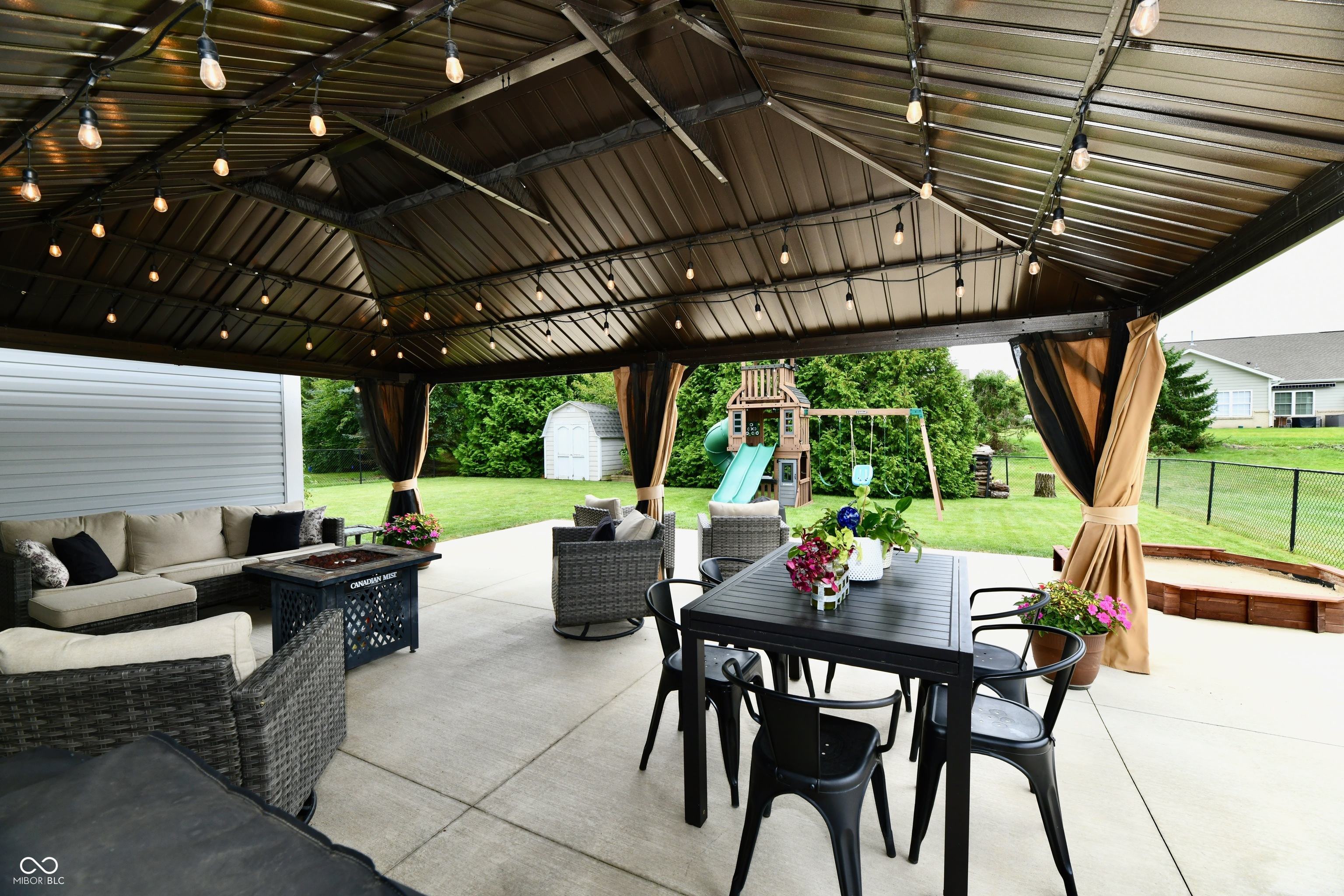 Covered backyard patio with wicker seating, dining table, string lights, and a playset in the yard.