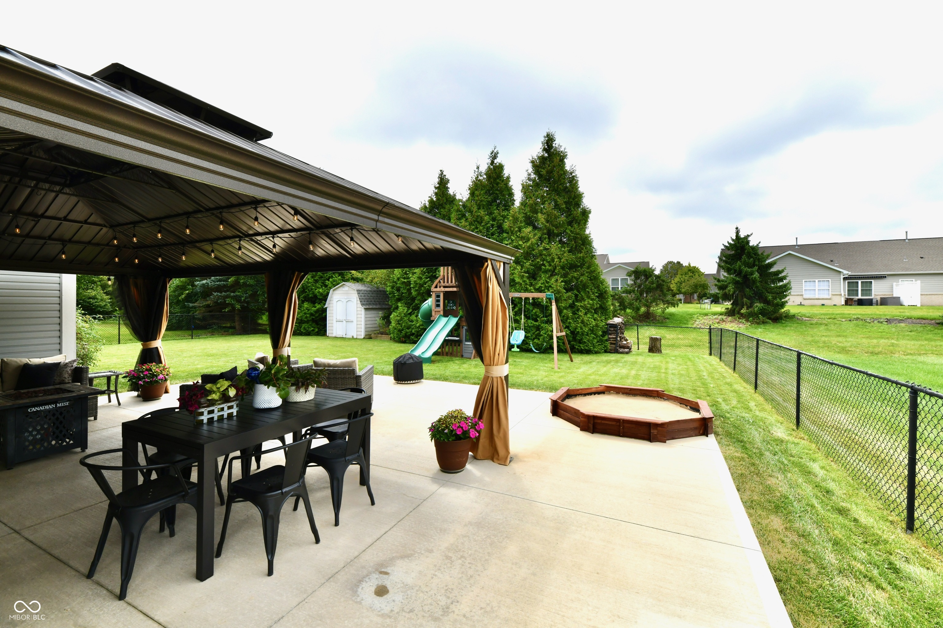 Covered patio with outdoor dining furniture overlooking a fenced backyard, sandbox, and children’s playset.