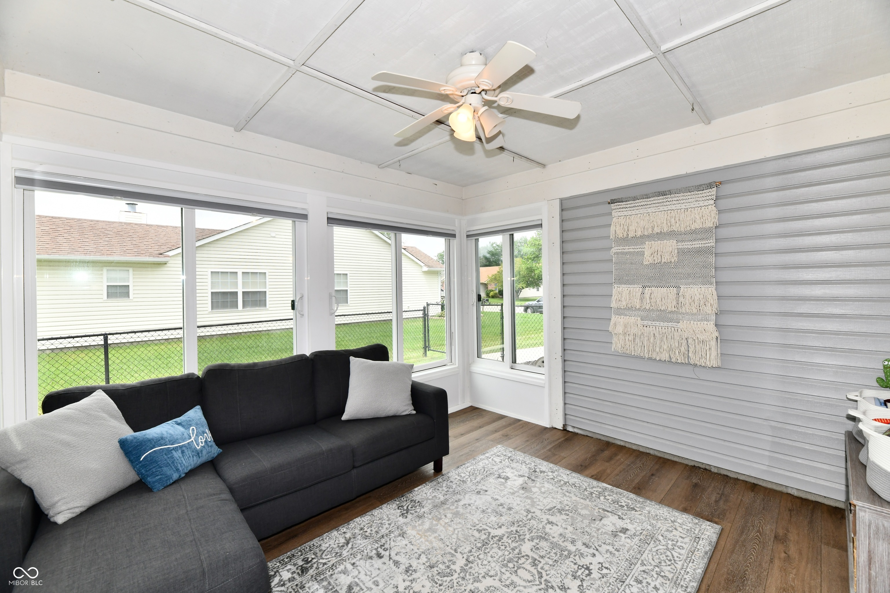 Furnished sunroom with a dark sectional, large windows, gray paneled wall, and ceiling fan.