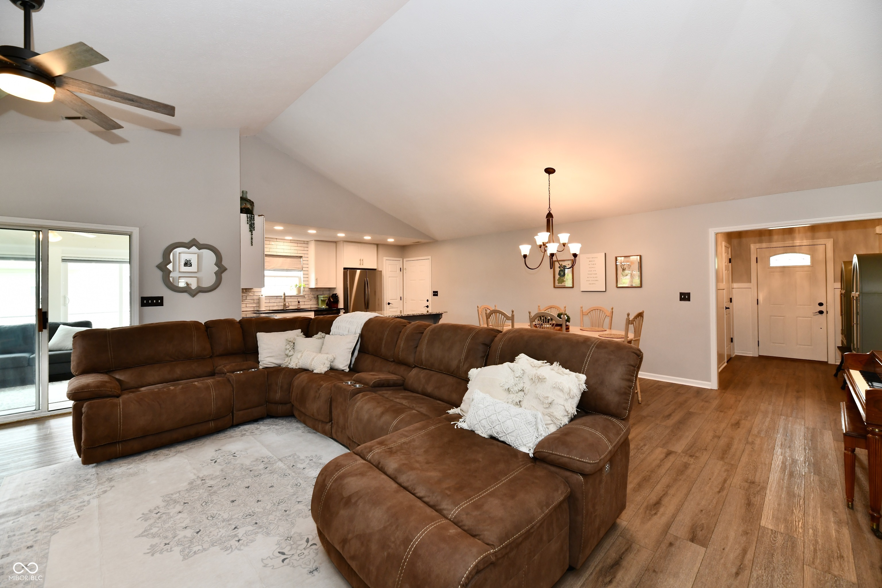 Open-plan living room with a large brown sectional, vaulted ceiling, and dining area beside the kitchen.