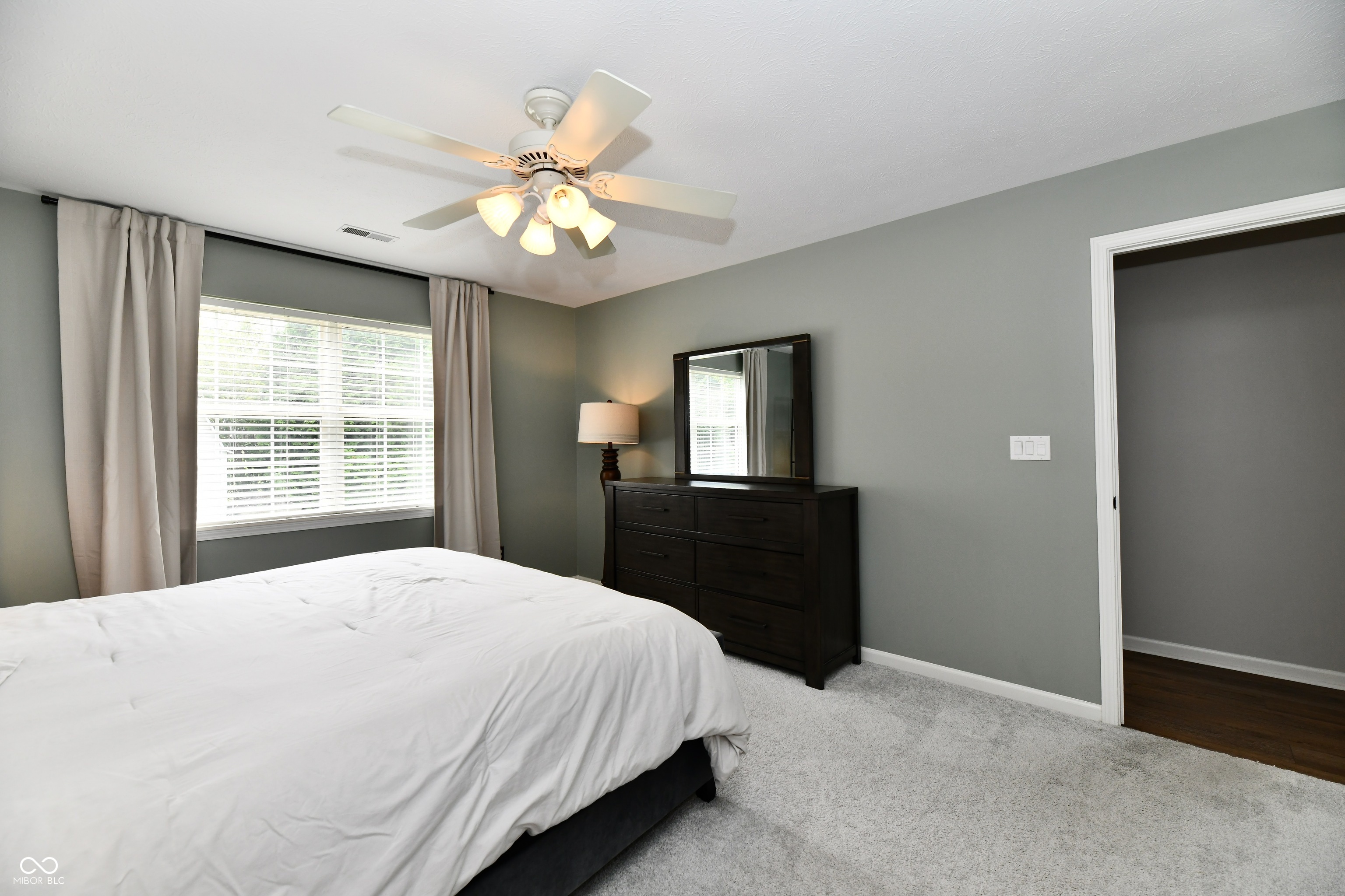 Furnished bedroom with white bedding, a bright curtained window, and a dark dresser and mirror.
