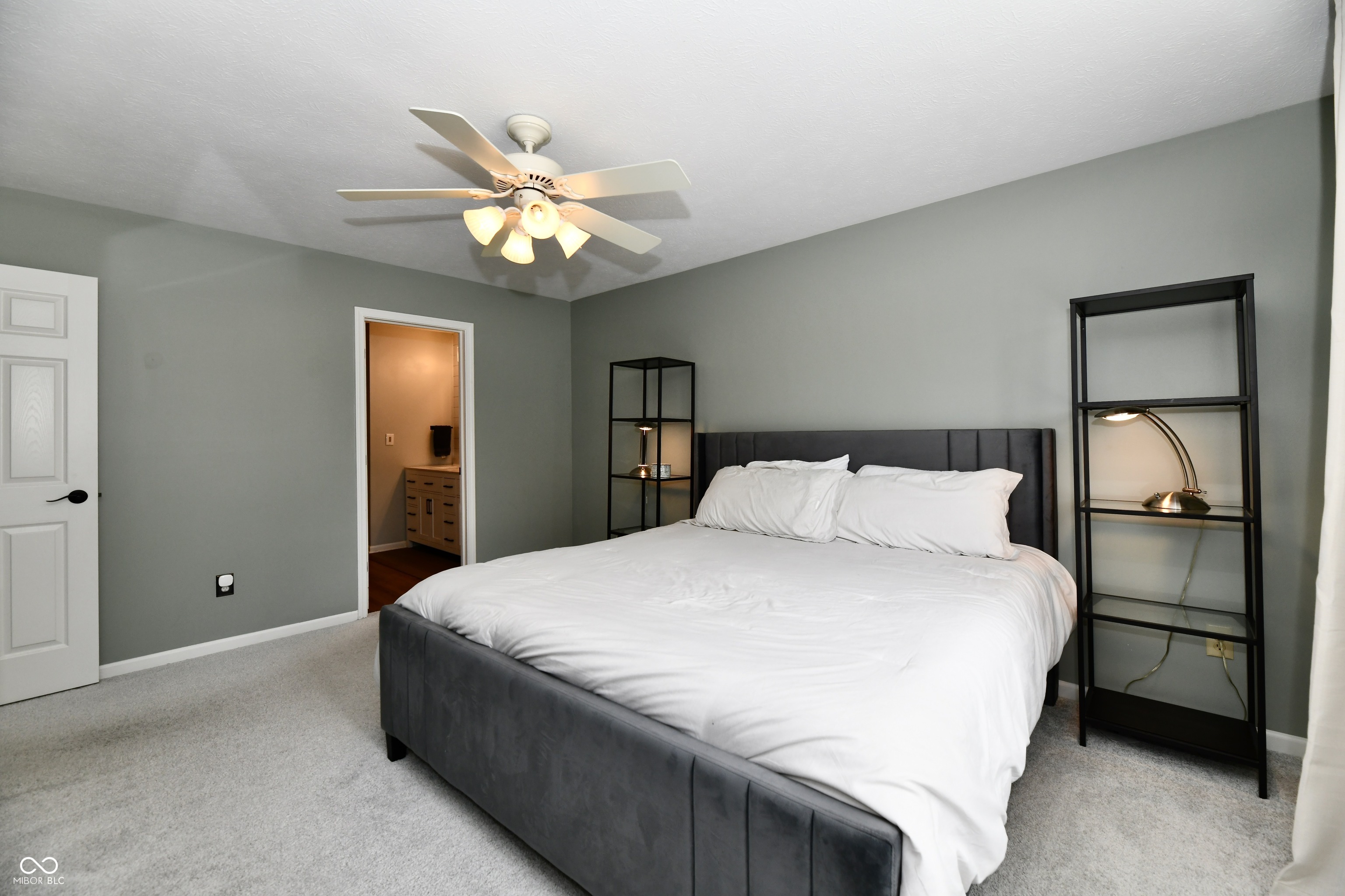 Furnished bedroom with a large white bed, gray walls, and black shelving beside the bed.