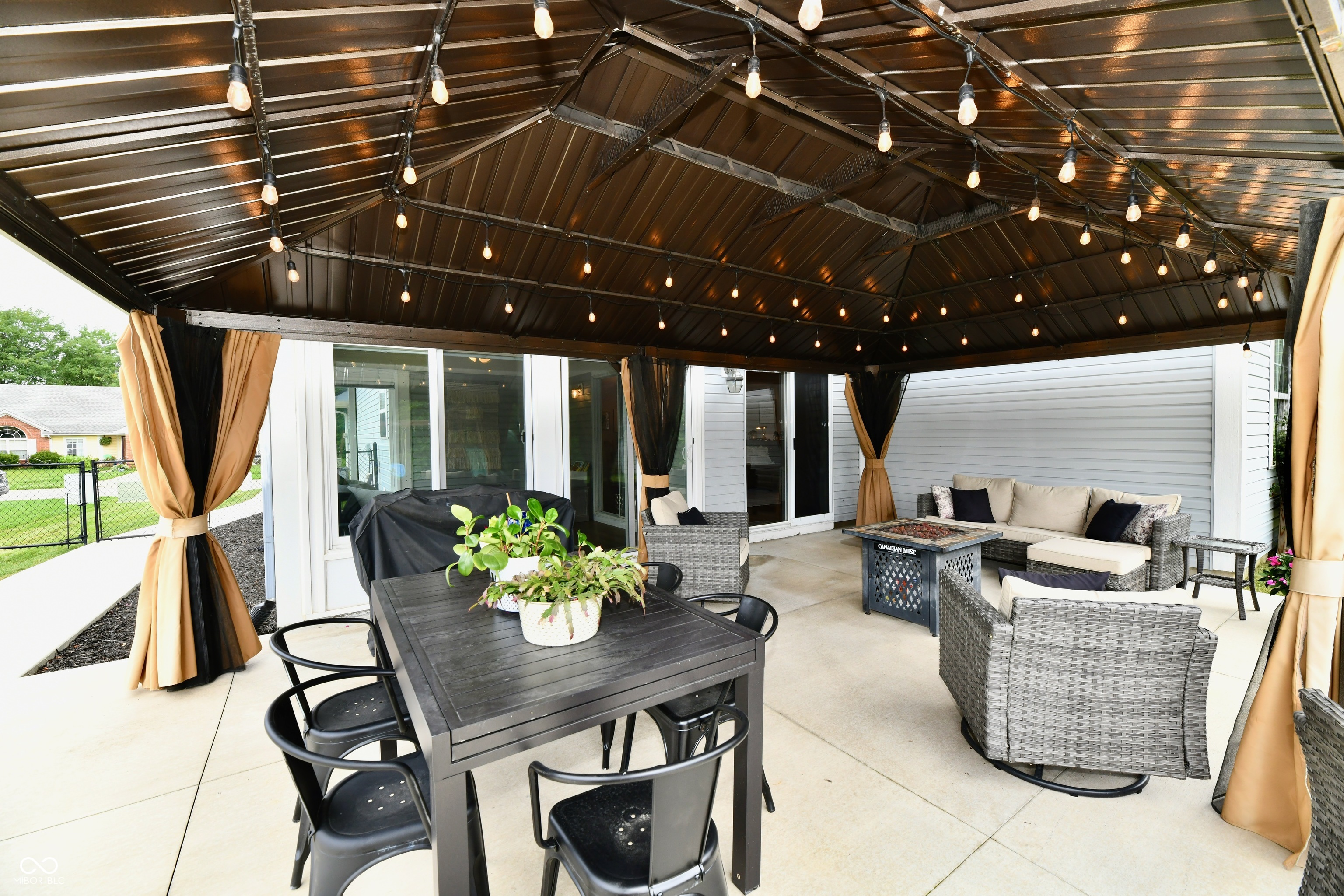 Covered patio with string lights, tan curtains, dining table, and wicker lounge seating.