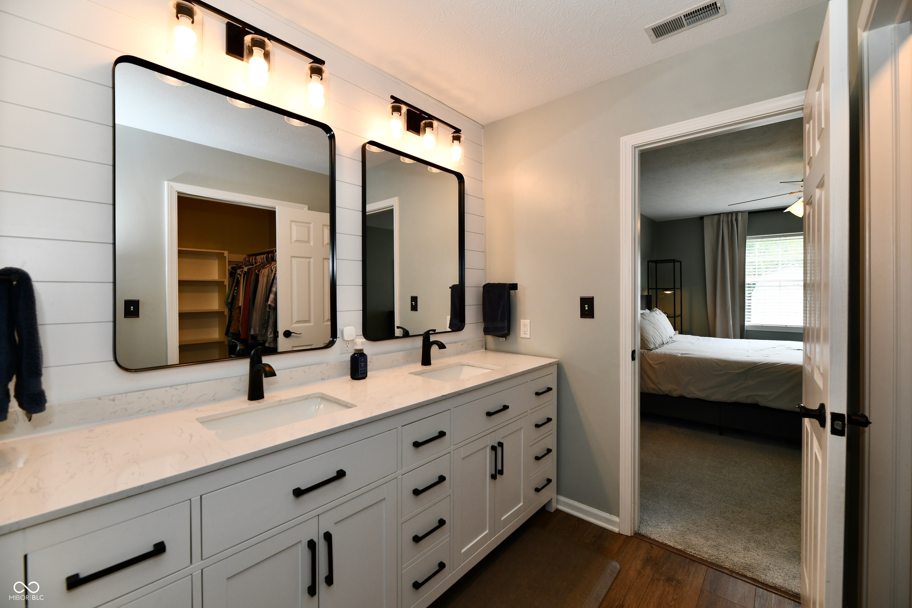 Bathroom with a white double vanity, marble-look counter, black fixtures, and a bedroom visible through the doorway.