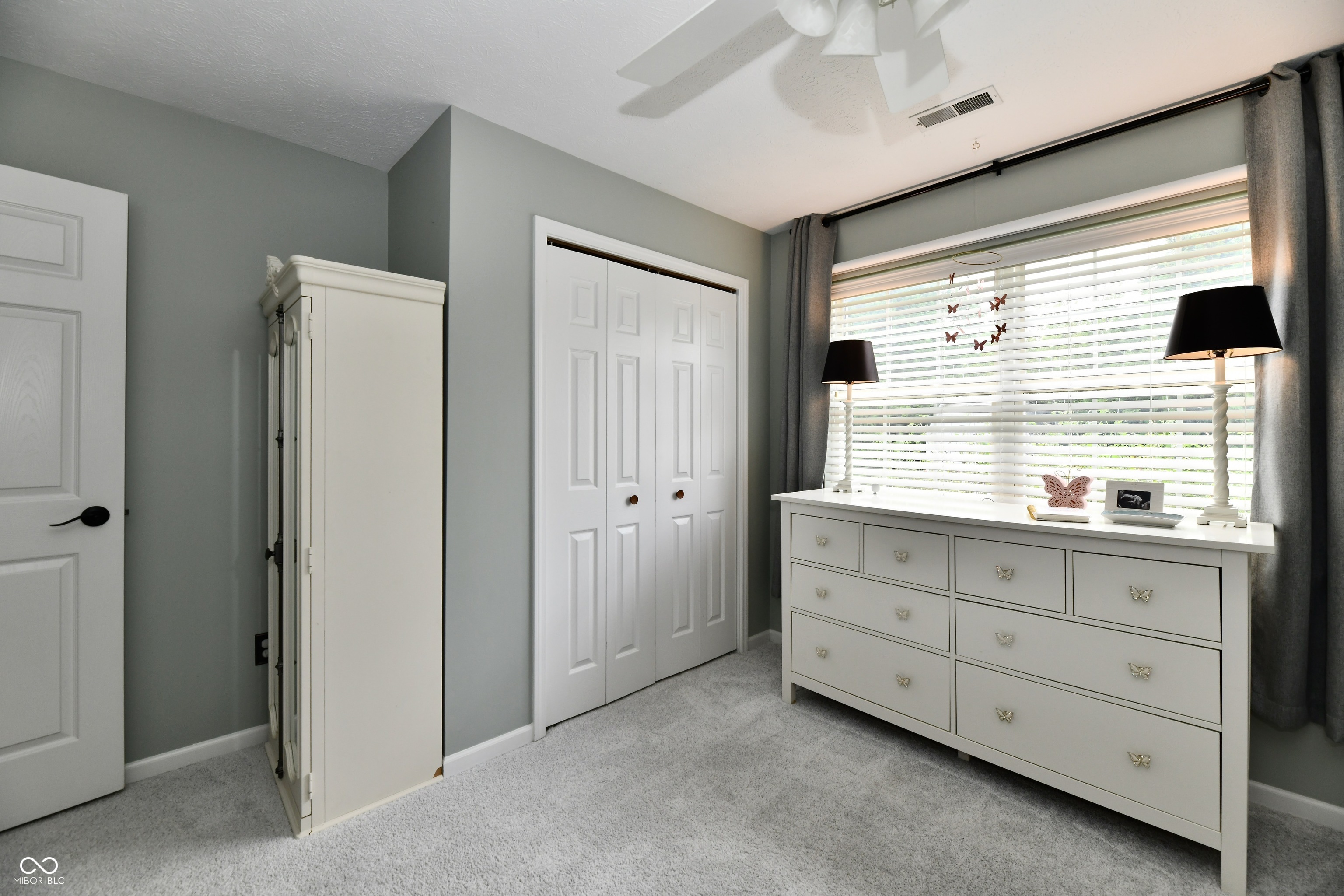 Bedroom with white wardrobe and dresser beneath a large window with blinds.