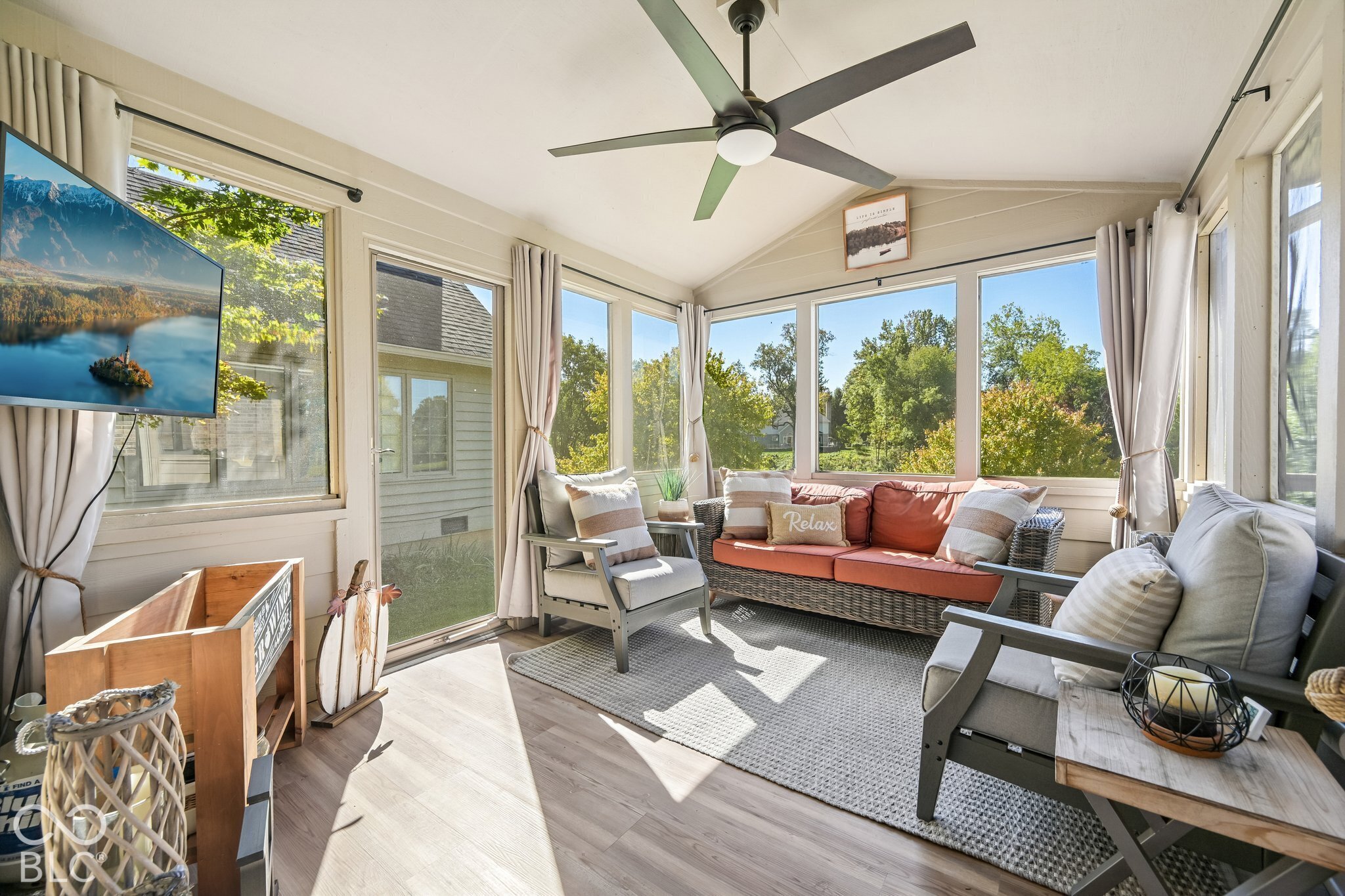 Sunroom with large windows, wicker sofa with coral cushions, gray armchairs, and a ceiling fan.