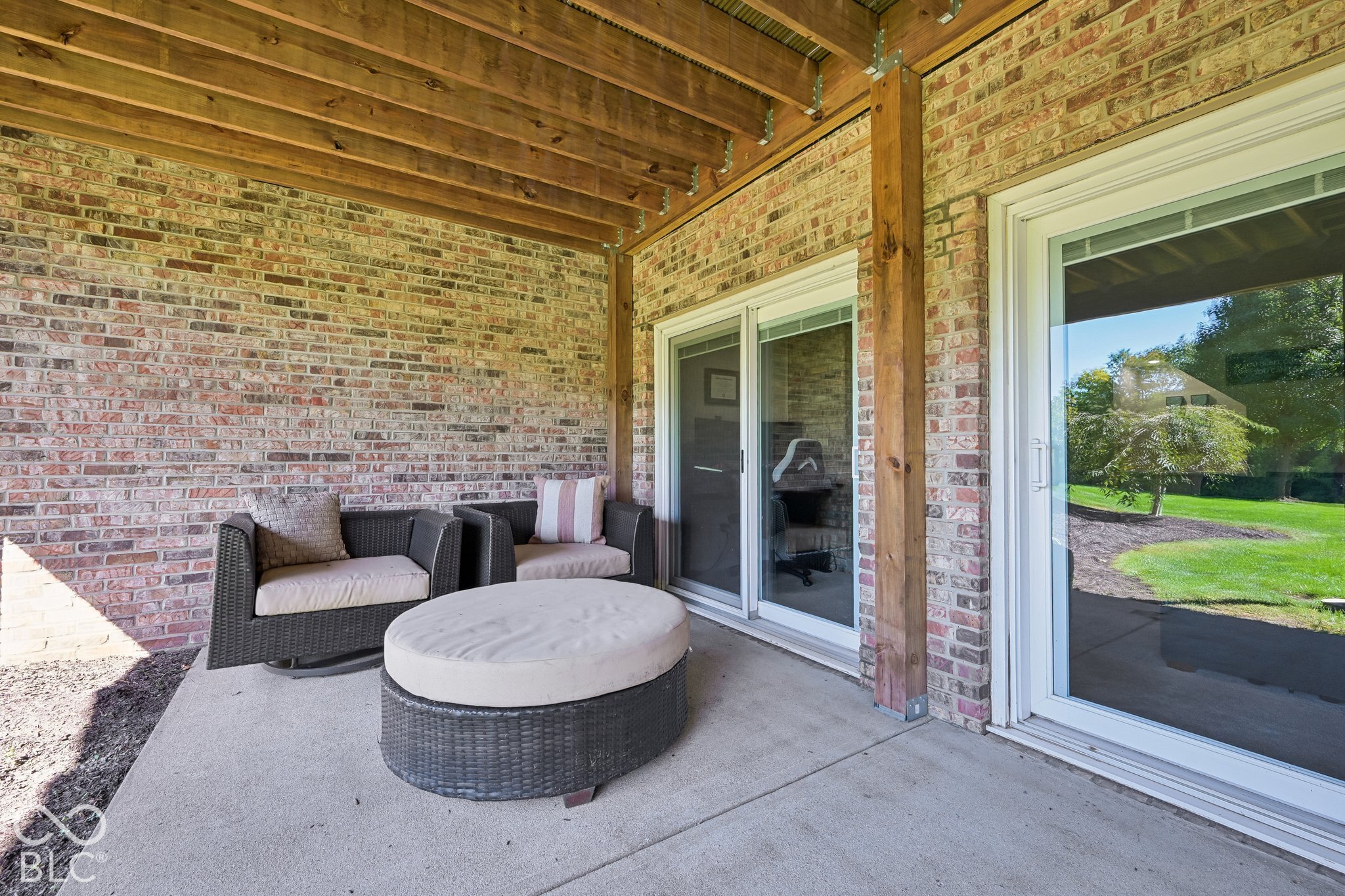 Covered brick patio with exposed wood beams, wicker seating, and sliding glass doors overlooking a lawn.
