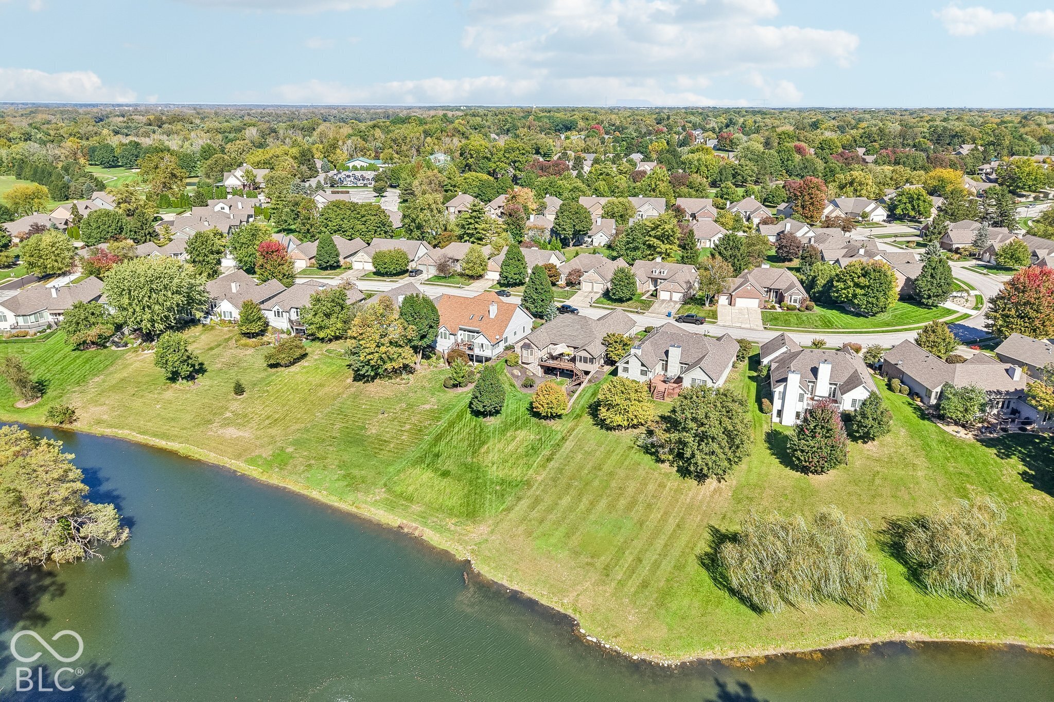 Aerial view of neighborhood homes and lawns along a pond, with autumn-colored trees.