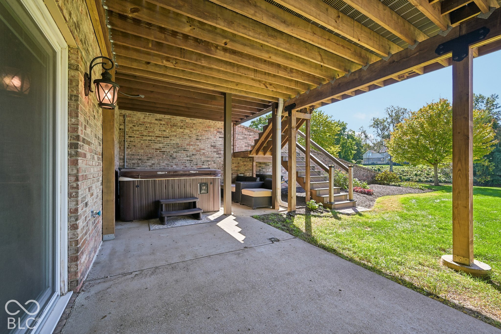 Covered brick patio with a hot tub, outdoor seating, and wooden stairs opening to a sunny lawn.