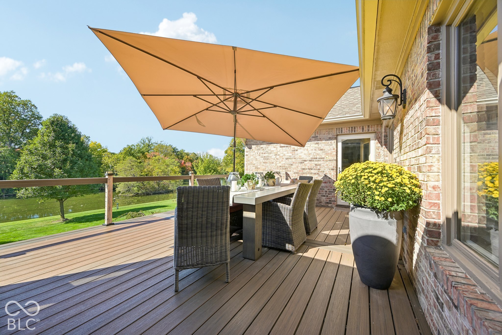 Wooden deck dining area shaded by a large tan umbrella, overlooking a lawn and trees.