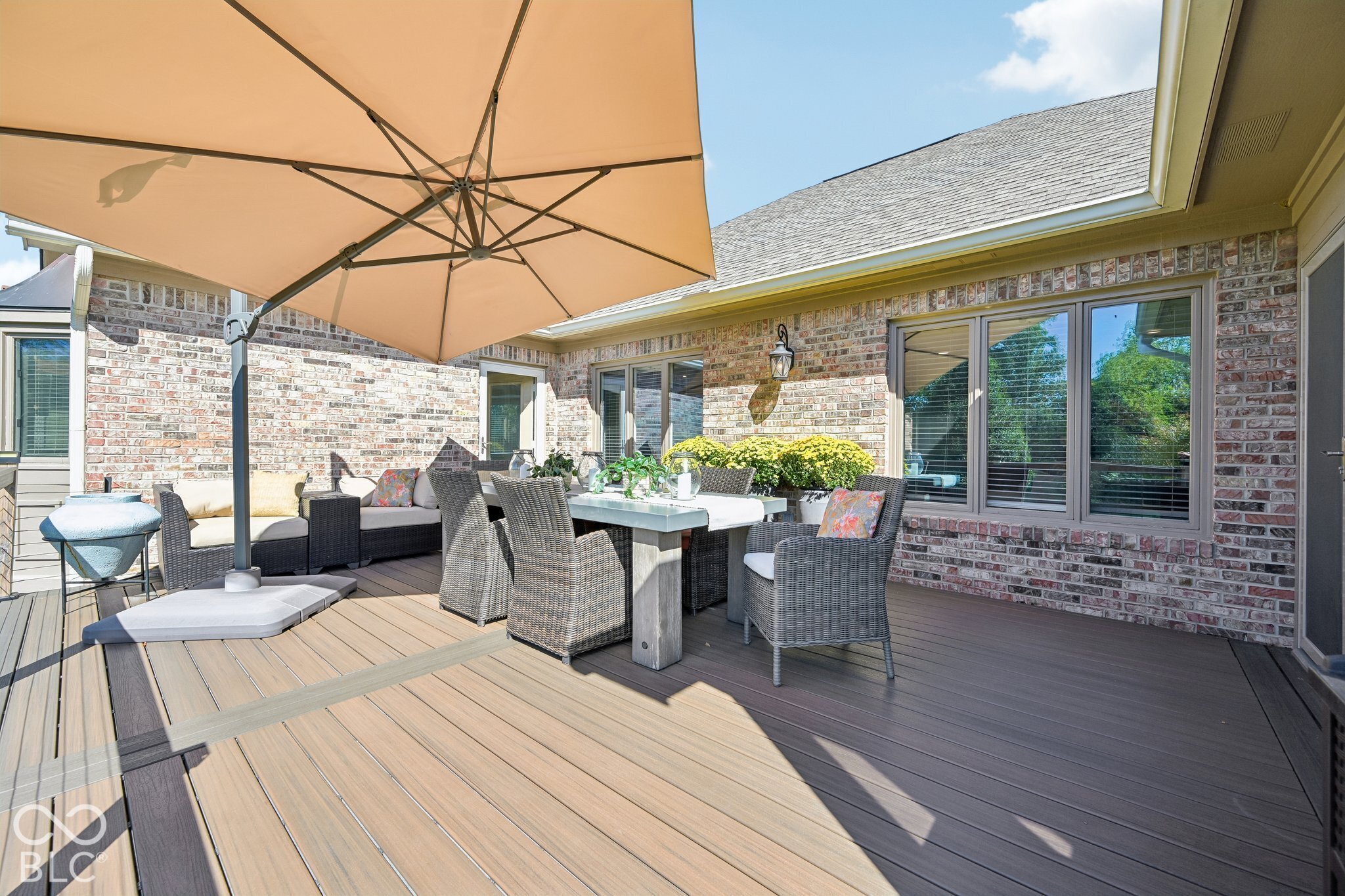 Outdoor deck with wicker dining and lounge seating beneath a large tan umbrella.