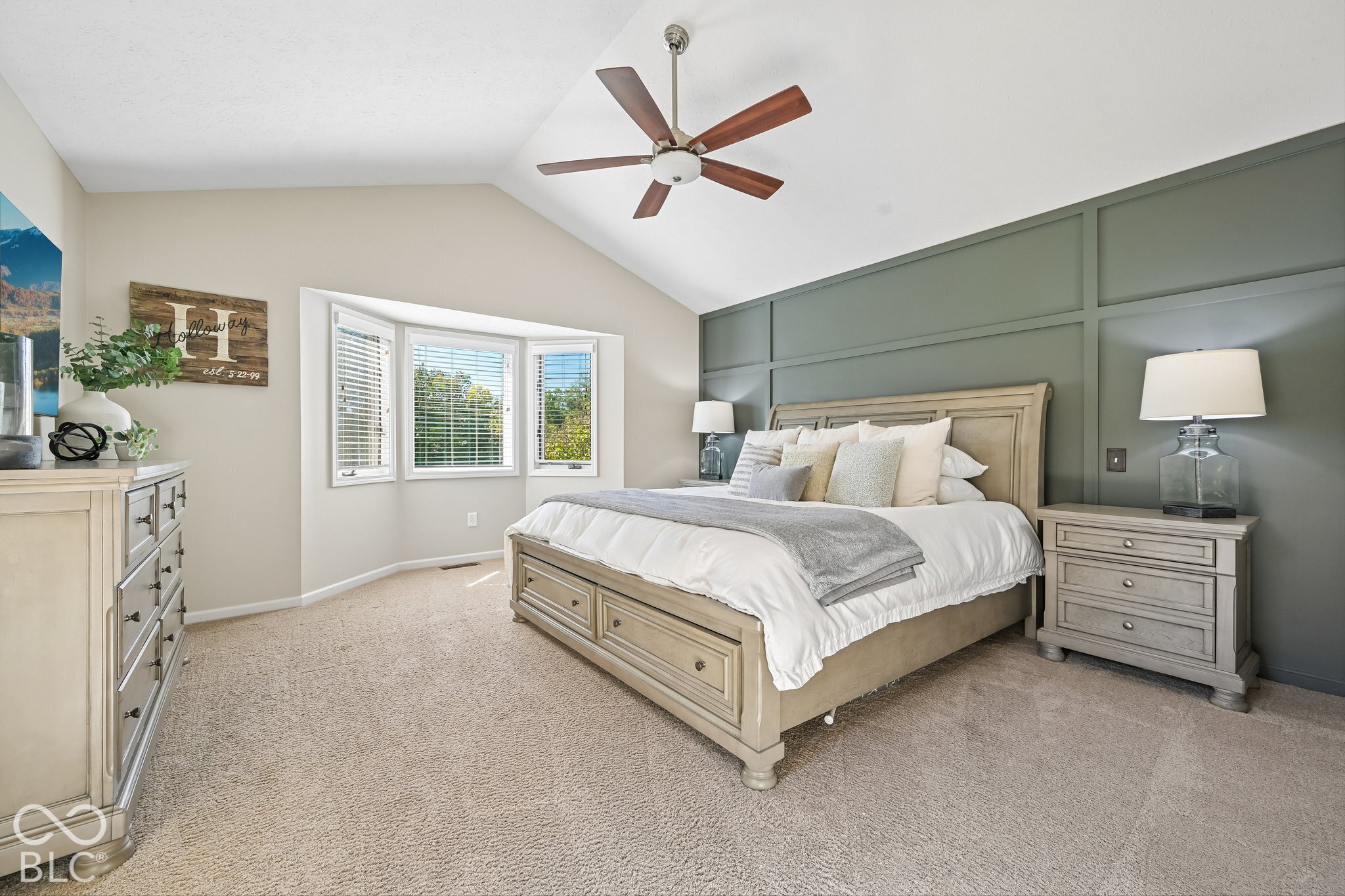 Staged bedroom with a beige bed, green paneled accent wall, bay window, and ceiling fan.