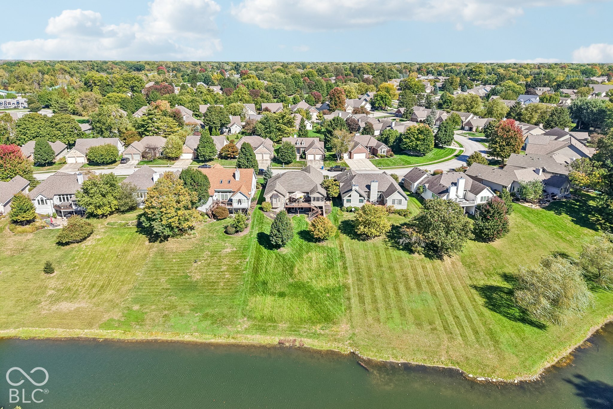 Aerial view of homes along a pond, with a broad grassy shoreline and mature trees.