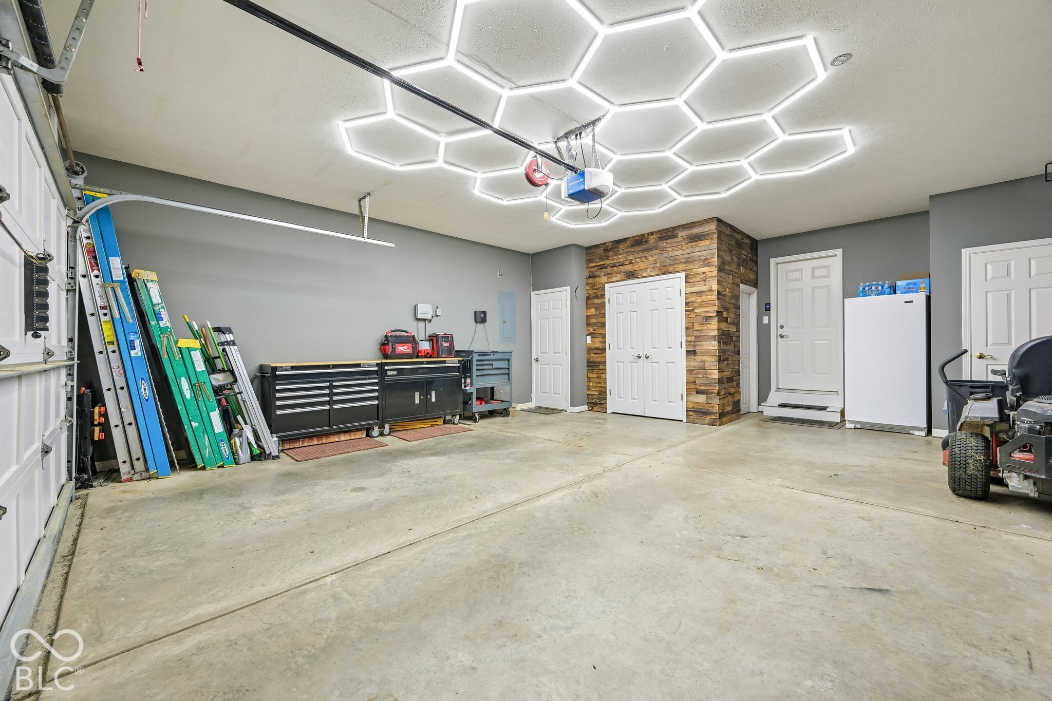Spacious garage with tool cabinets, ladders, a wood-plank accent wall, and bright hexagonal ceiling lights.