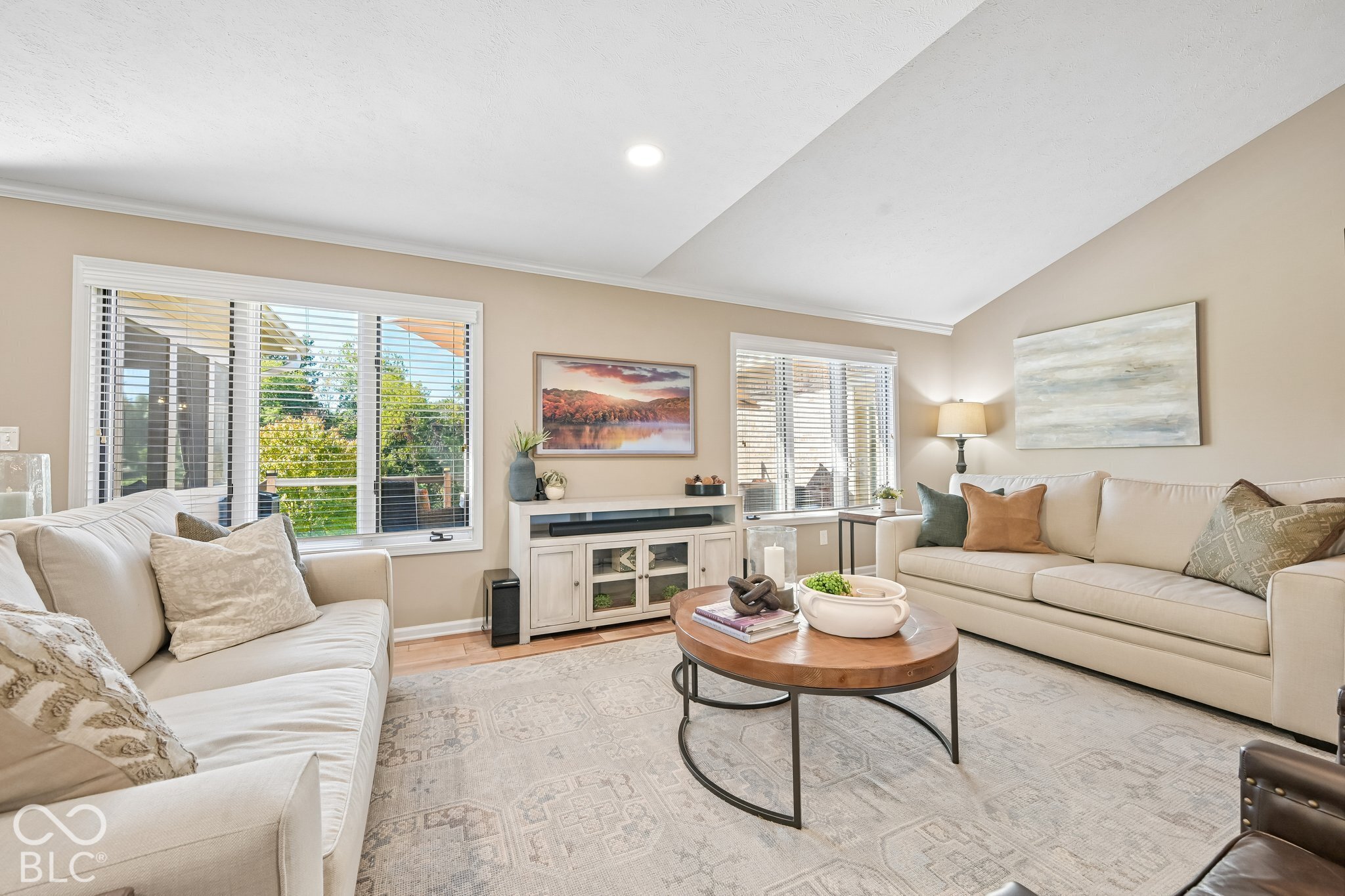 Furnished living room with cream sofas, a round wood coffee table, and large windows.