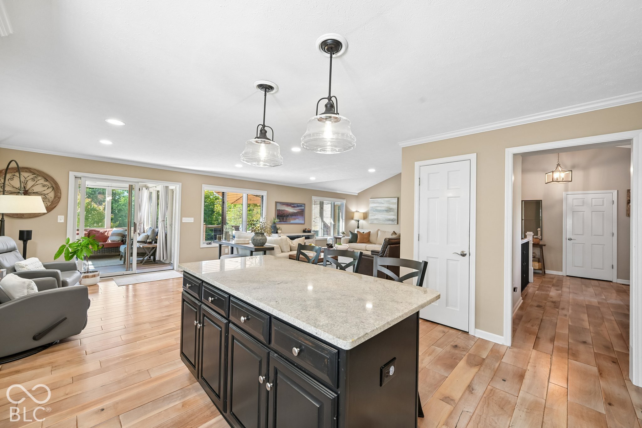 Open-plan kitchen with a large dark island, stone countertop, glass pendants, and adjoining living room.