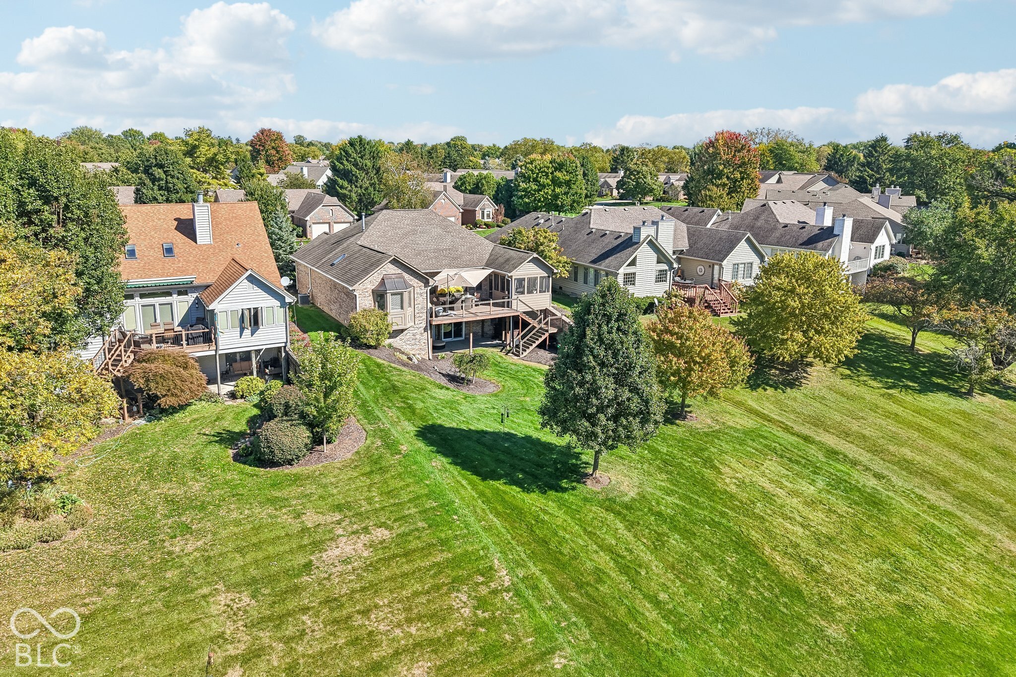 Aerial view of a brick home’s rear deck, broad lawn, mature trees, and neighboring houses.