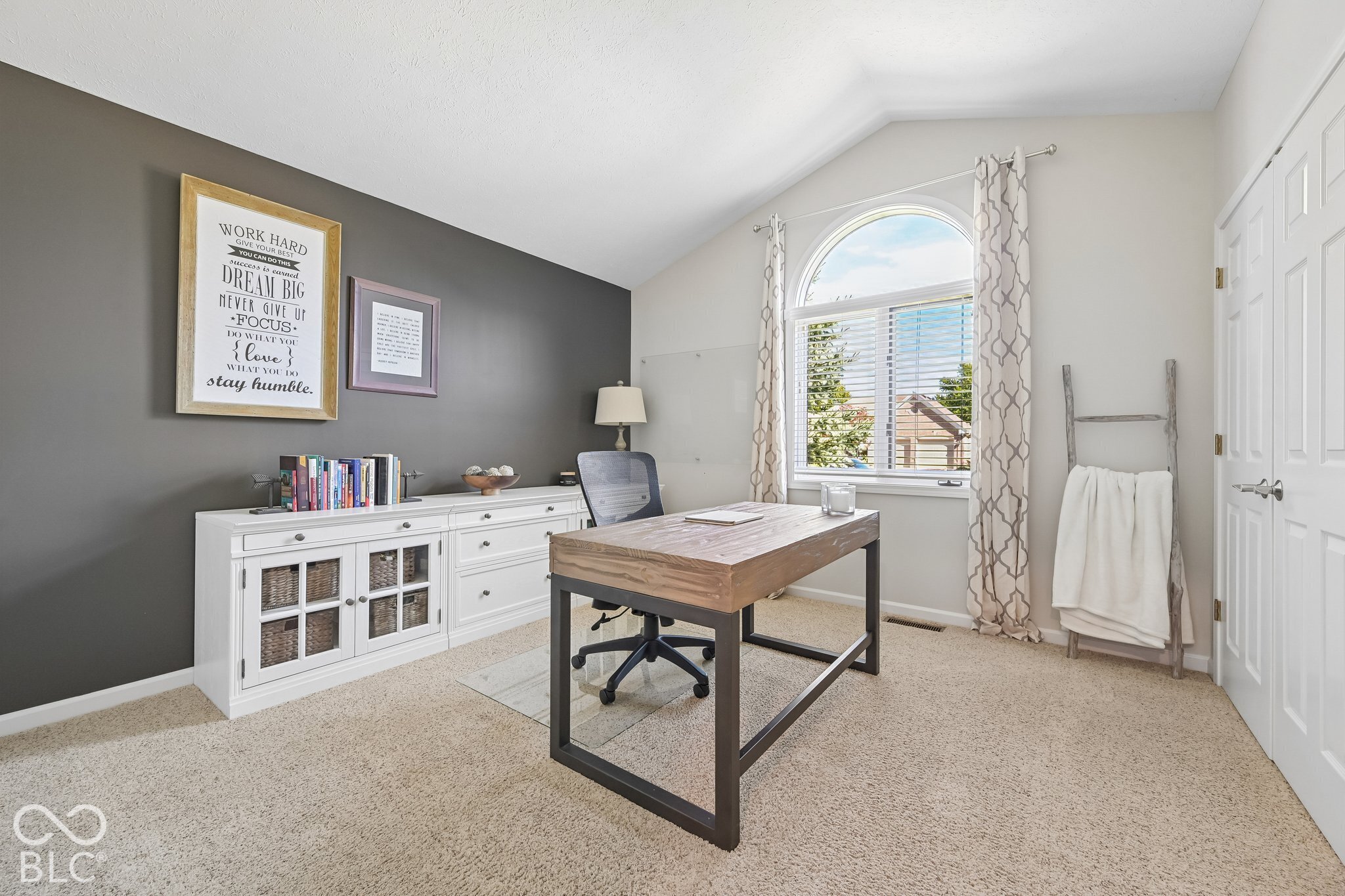 Home office with a wooden desk, white storage cabinets, and an arched window with patterned curtains.