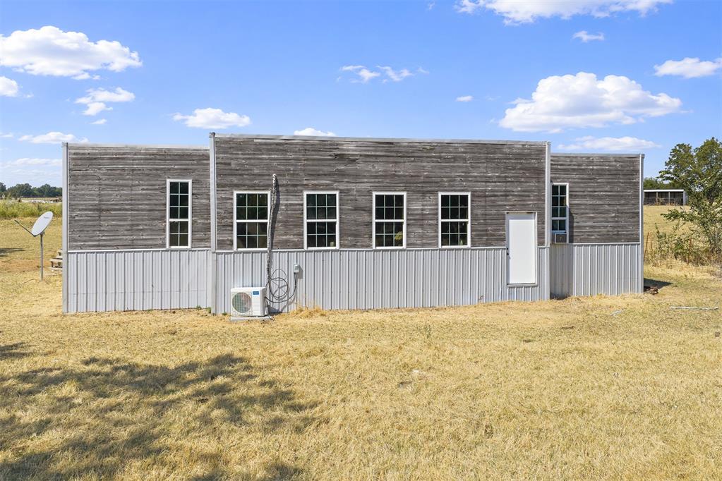 Long gray-sided rural building with tall windows, a white door, and dry grass in front.