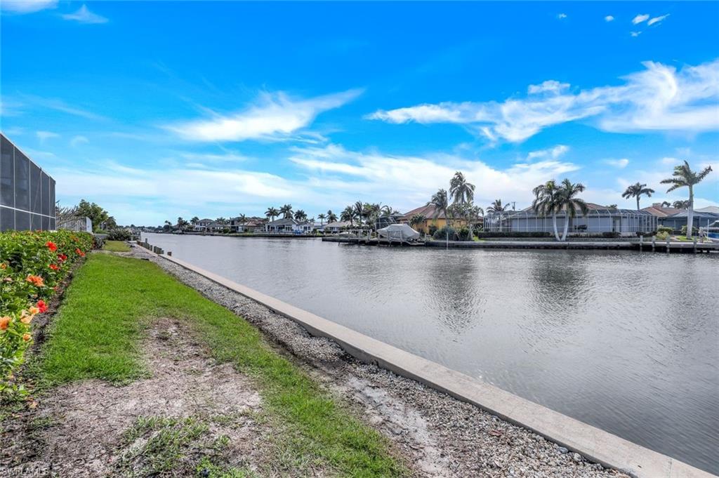 Residential canal with a flower-lined grassy bank and waterfront homes across the water.