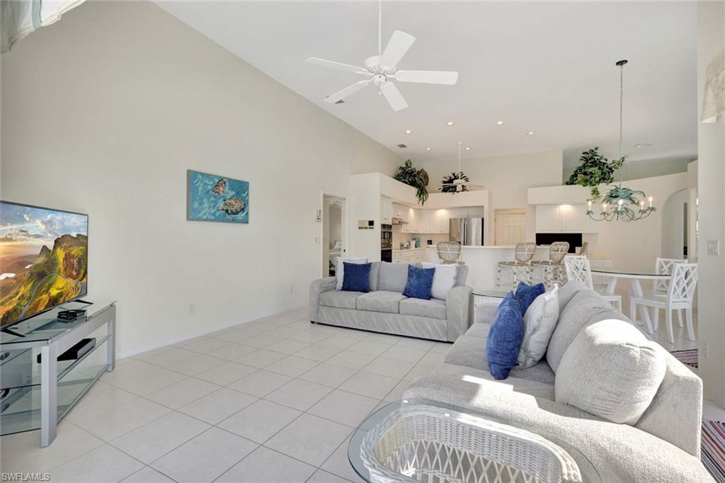 Living room with vaulted ceiling, white tile floors, blue-accented sofas, and an open kitchen beyond.