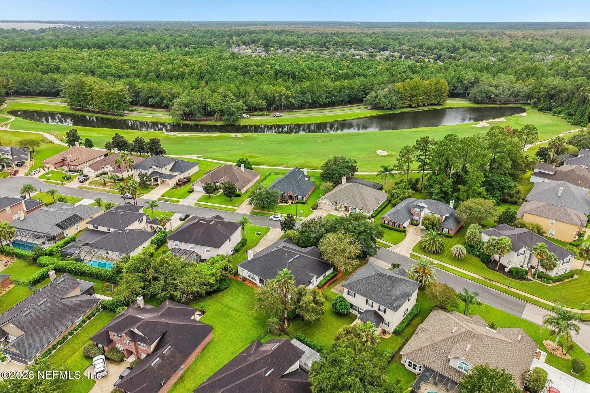 Aerial view of homes beside a golf course and pond, with wooded land in the distance.