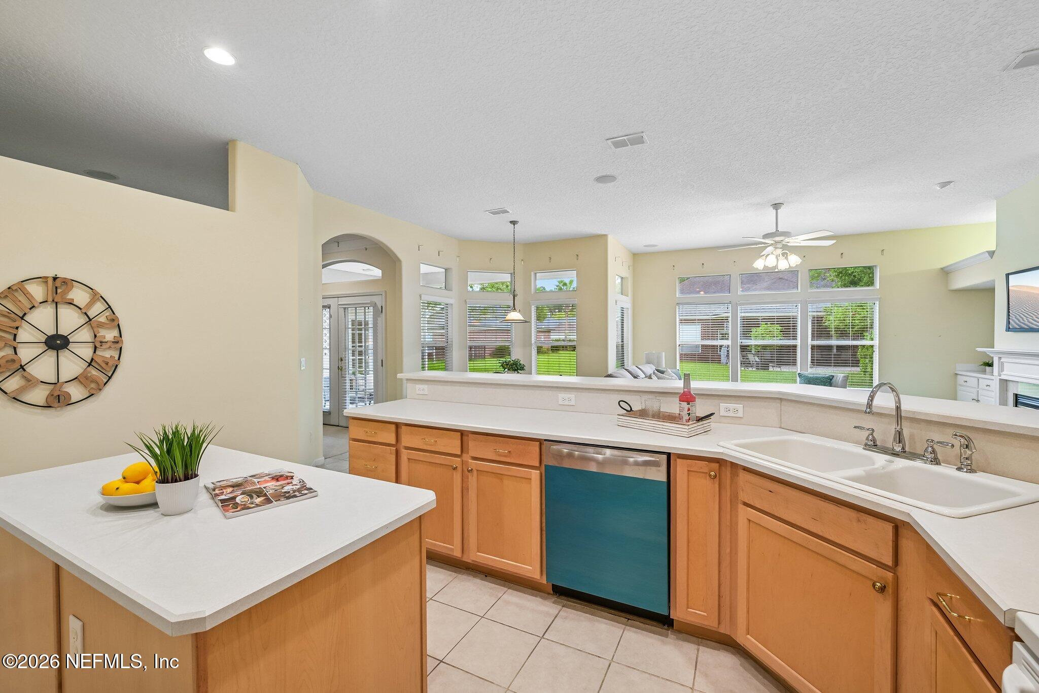 Open kitchen with wood cabinets, white counters, teal-front dishwasher, and windows overlooking a green yard.