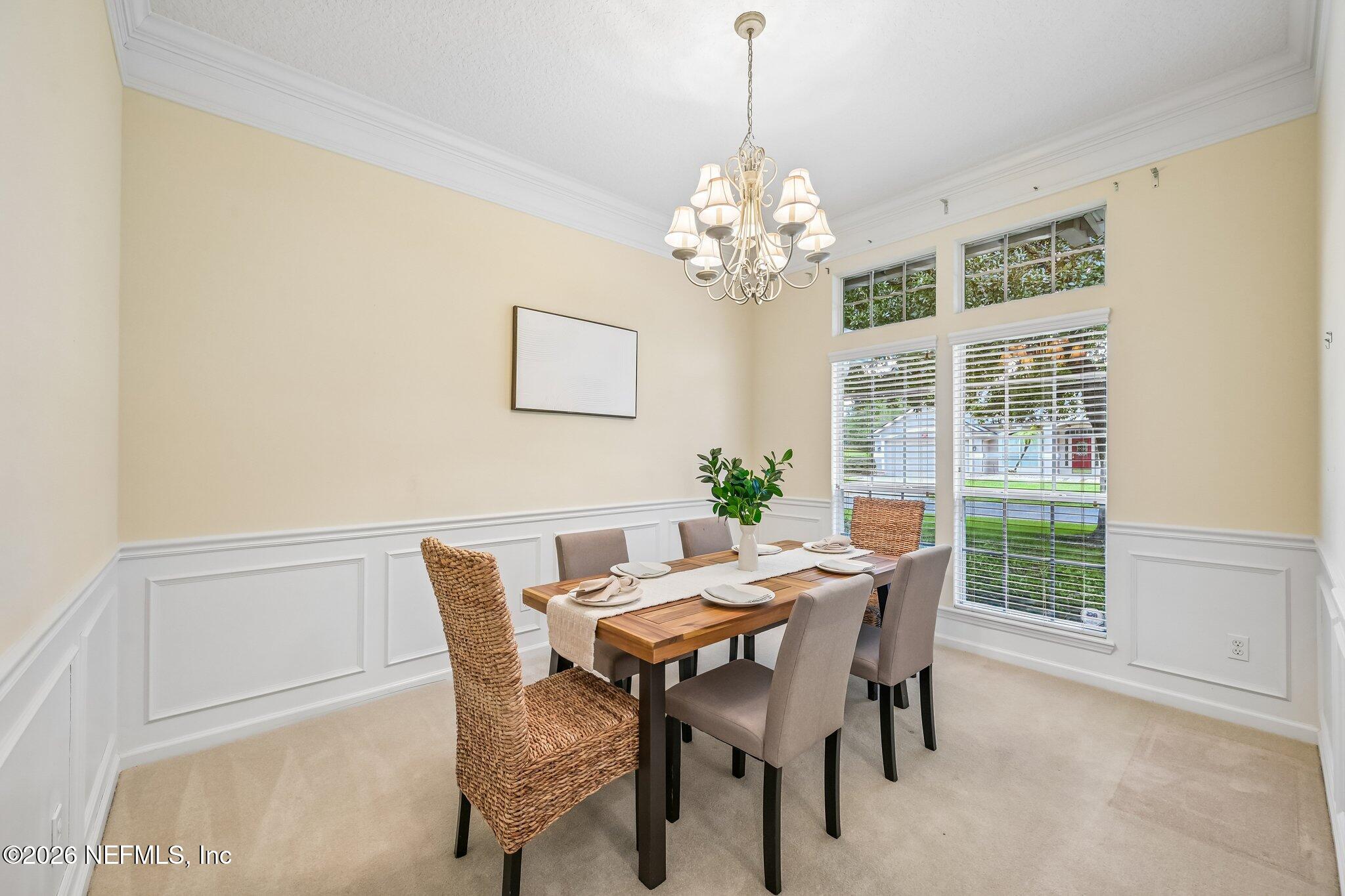 Dining room with a set wooden table, chandelier, white wainscoting, and tall windows.