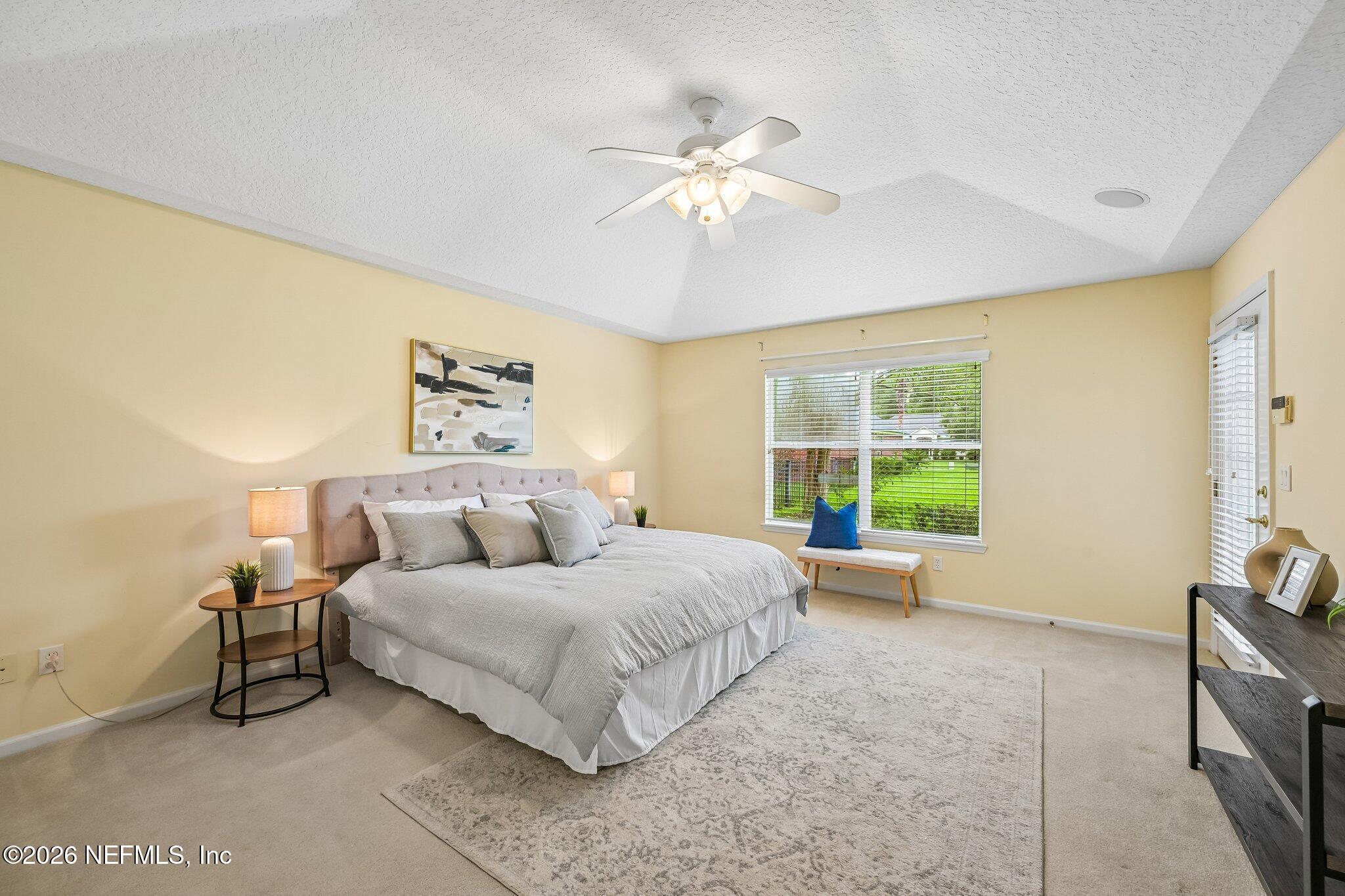 Staged bedroom with a large gray-and-white bed, ceiling fan, and window overlooking a green lawn.