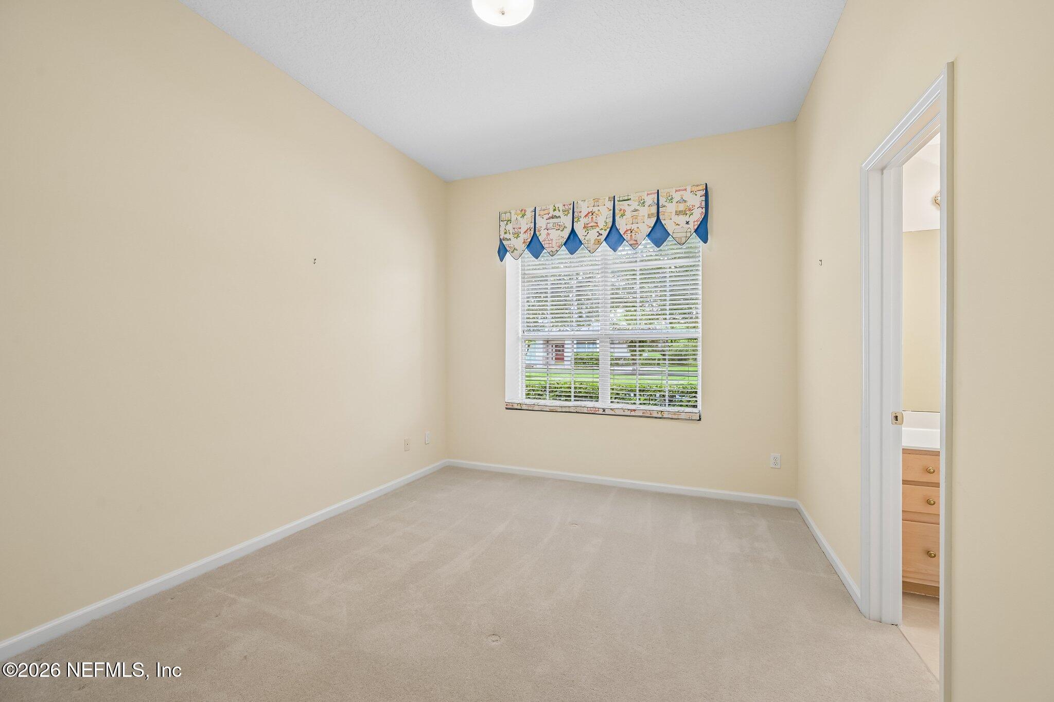 Empty bedroom with beige walls, carpet, and a window with white blinds and a patterned valance.