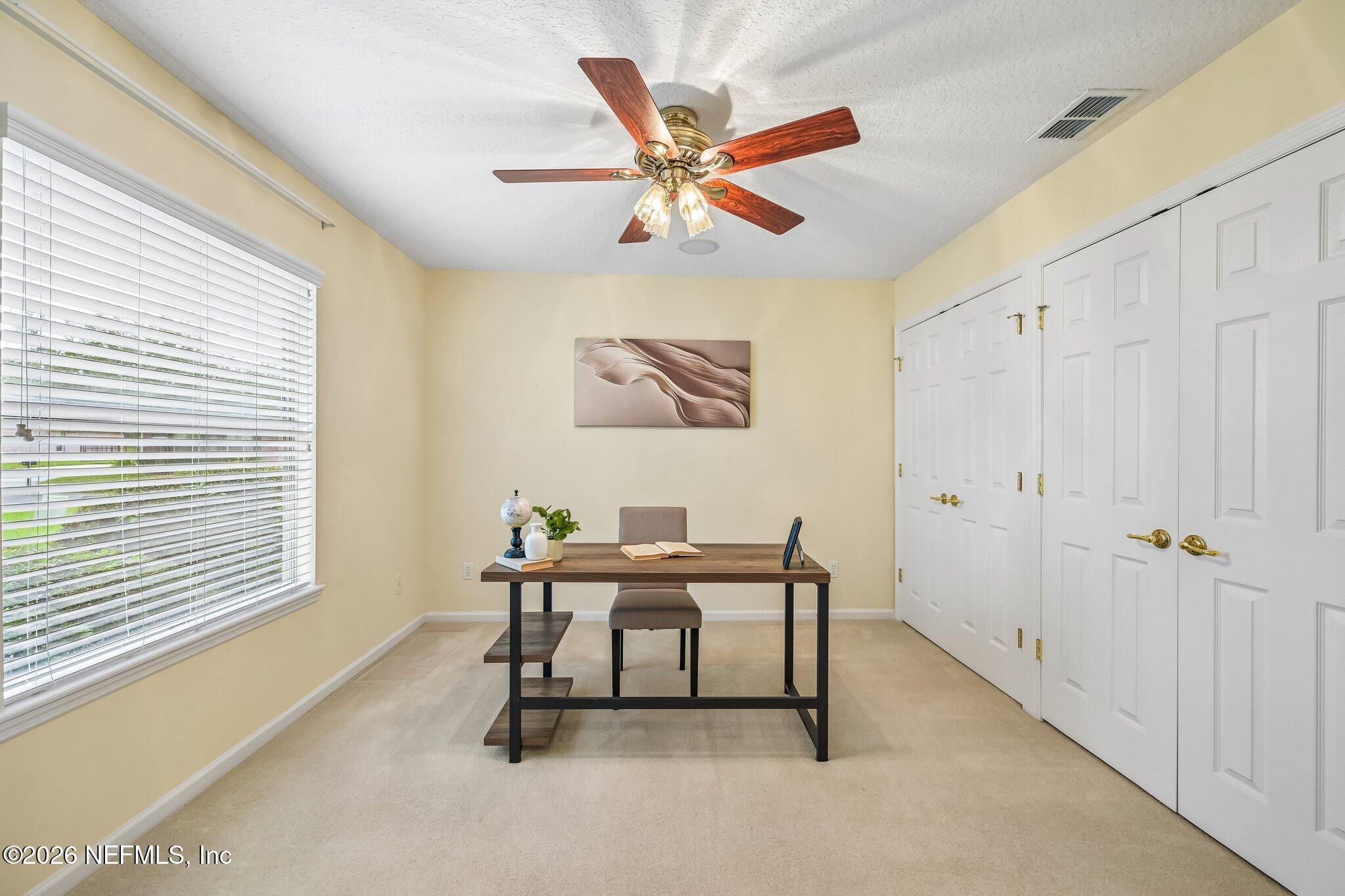 Staged home office with a desk beneath abstract wall art, a wide window, and white closet doors.