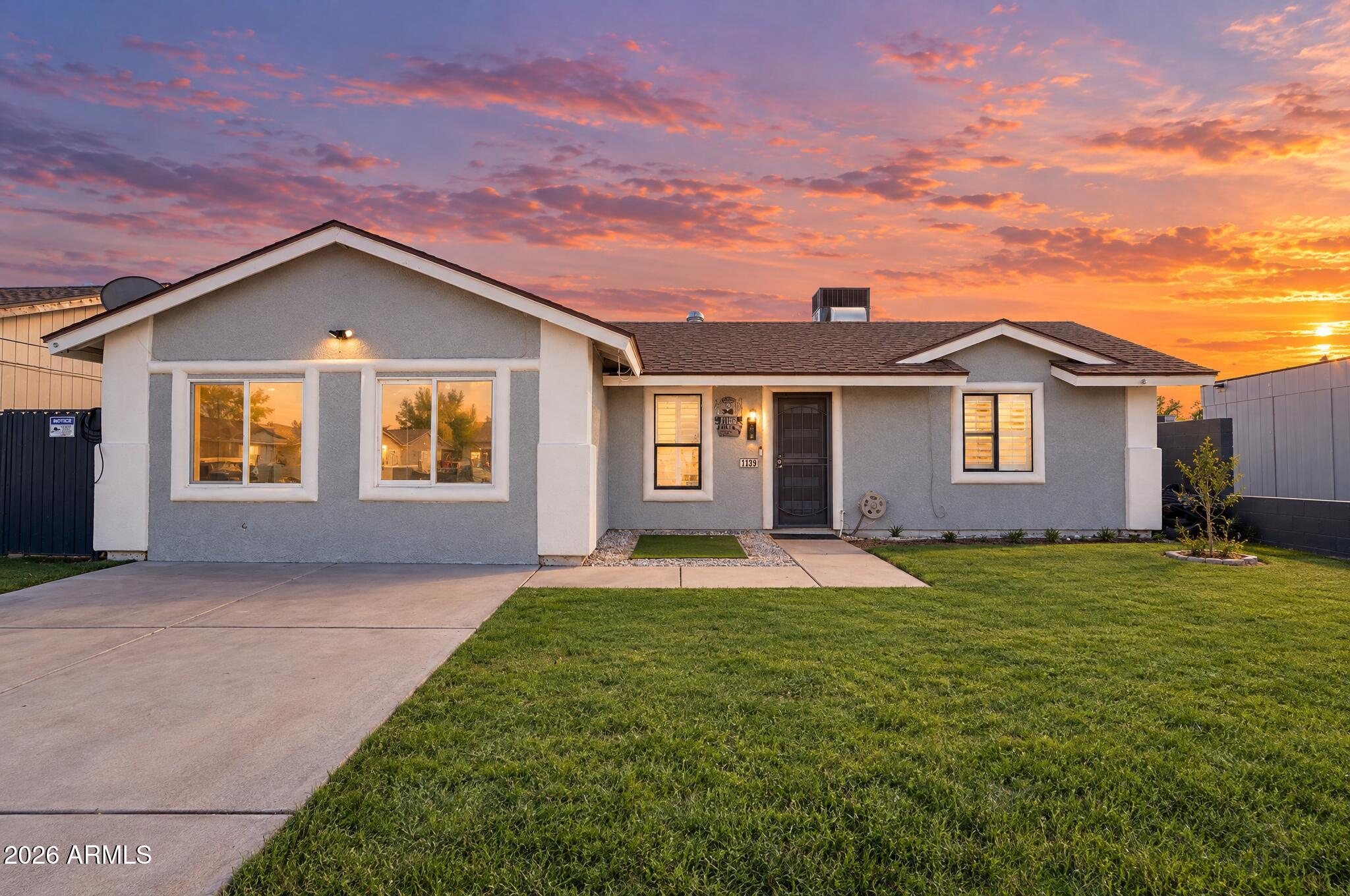 Single-story gray stucco house with a green lawn beneath a vivid sunset.
