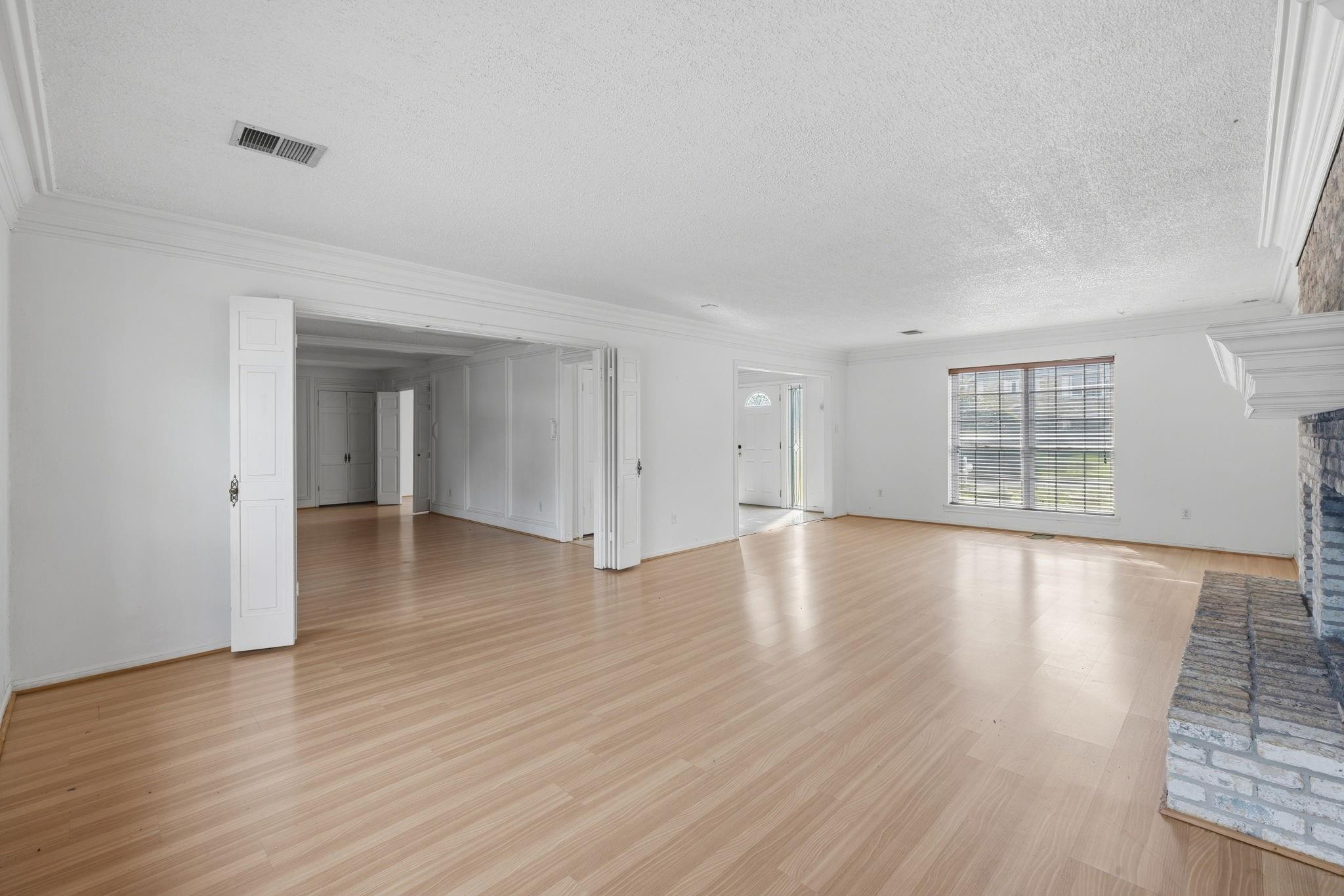 Empty living room with light wood floors, open double doors, a large blinded window, and a stone fireplace.
