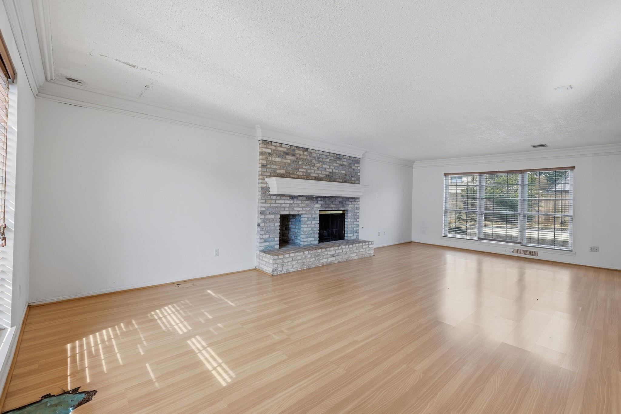 Vacant living room with a brick fireplace, light wood flooring, and wide windows with blinds.