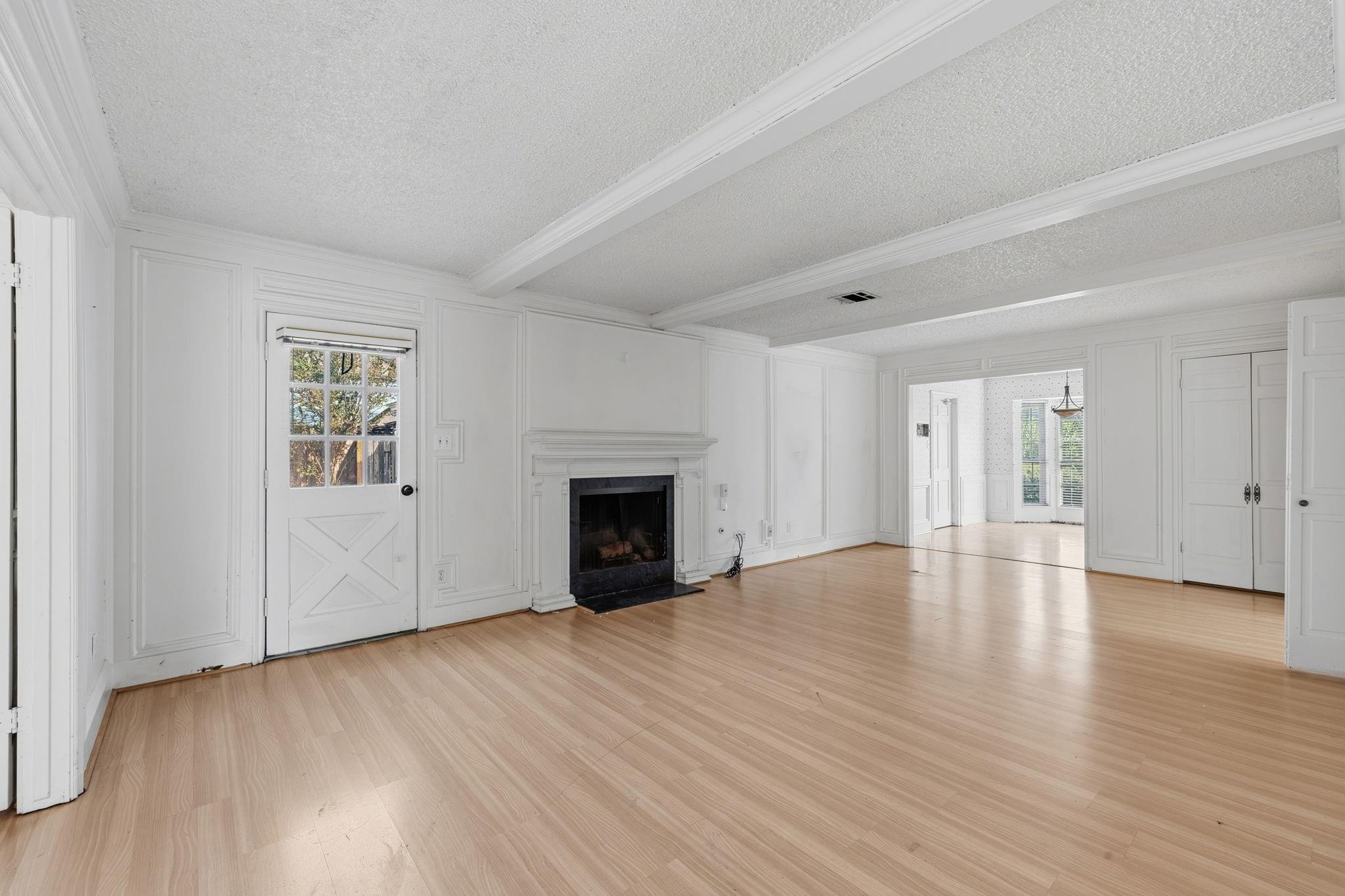 Empty living room with a fireplace, white paneled walls, light wood floors, and a beamed ceiling.