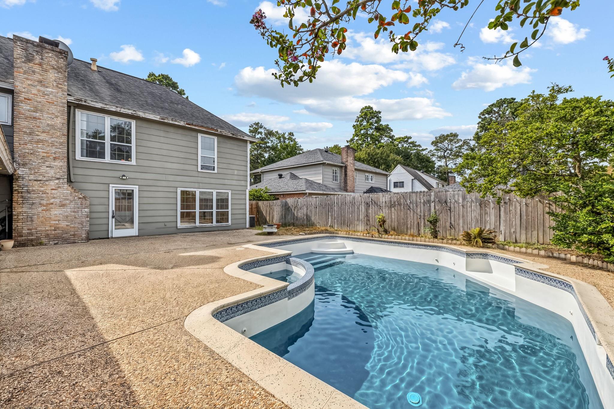 Backyard swimming pool beside a two-story home, with a broad patio and wood privacy fence.