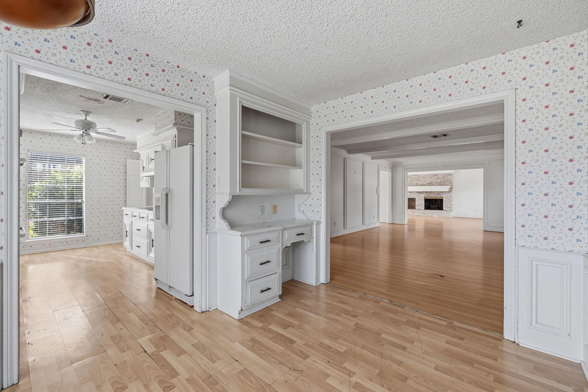 Vacant kitchen with floral wallpaper and built-in cabinets, opening toward a room with a brick fireplace.