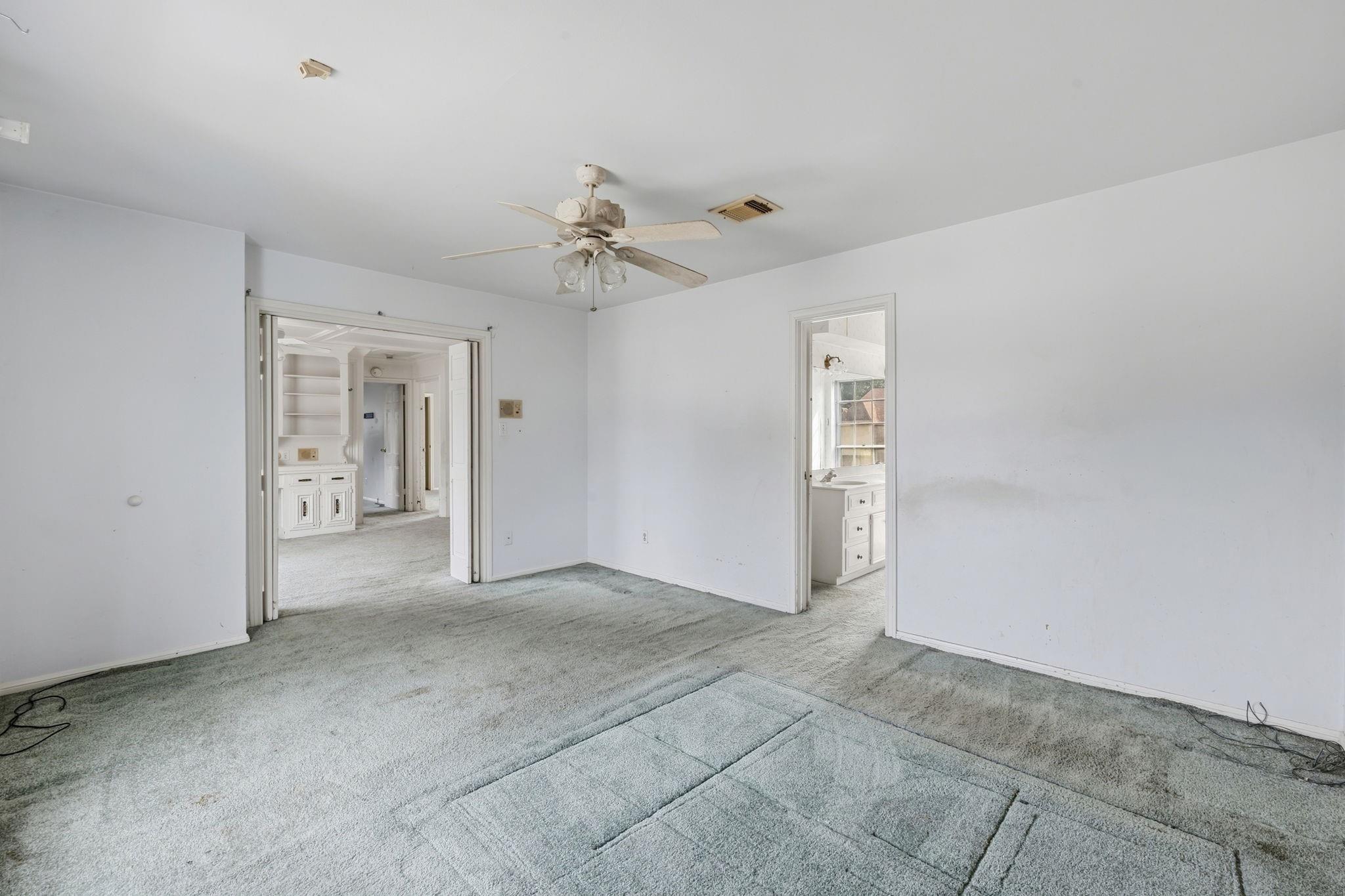 Empty carpeted bedroom with a ceiling fan, built-in cabinetry, and a bathroom vanity visible through open doorways.