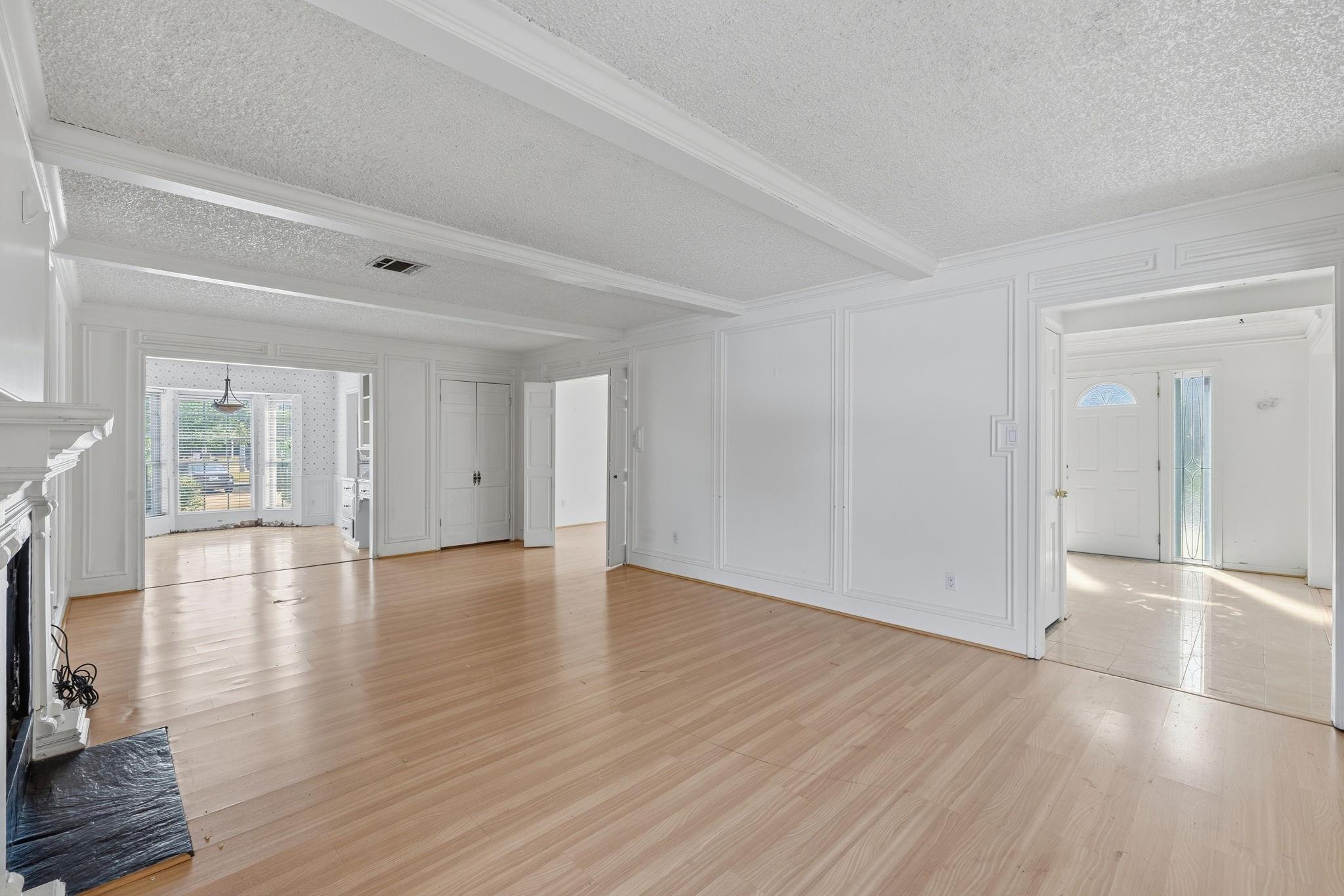 Vacant living room with white wall paneling, a beamed ceiling, light wood floors, and a fireplace.