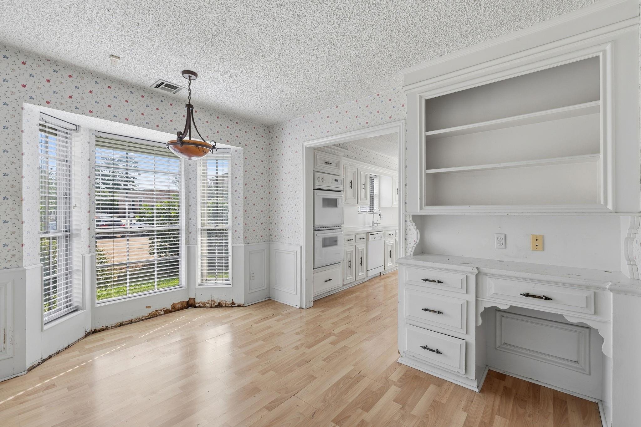 Vacant dining area with a built-in white buffet, floral wallpaper, and peeling trim beneath large windows.