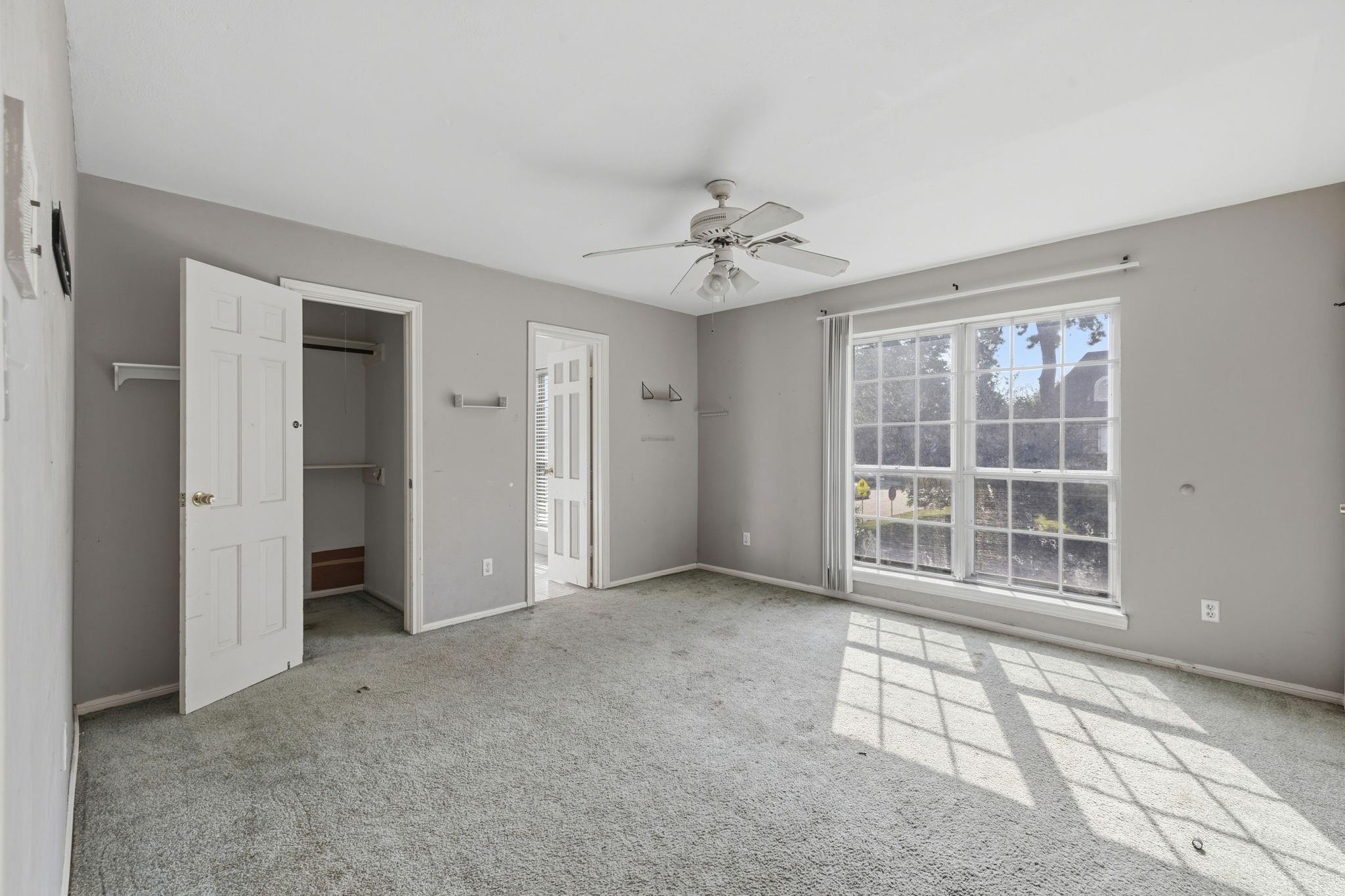 Empty carpeted bedroom with an open closet, ceiling fan, and large sunlit window.