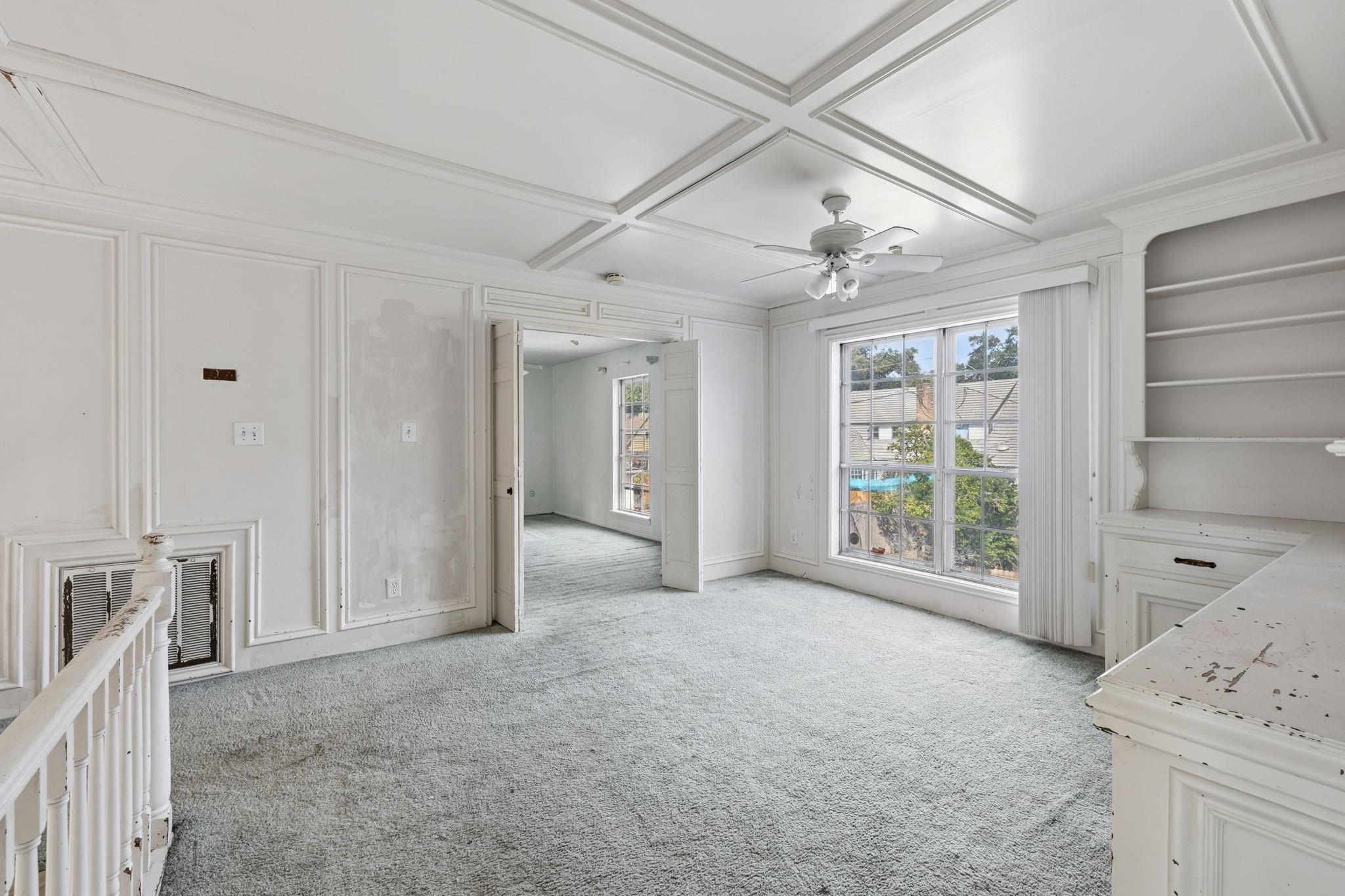 Empty upstairs room with a coffered ceiling, large window, built-in shelves, and doorway to another room.