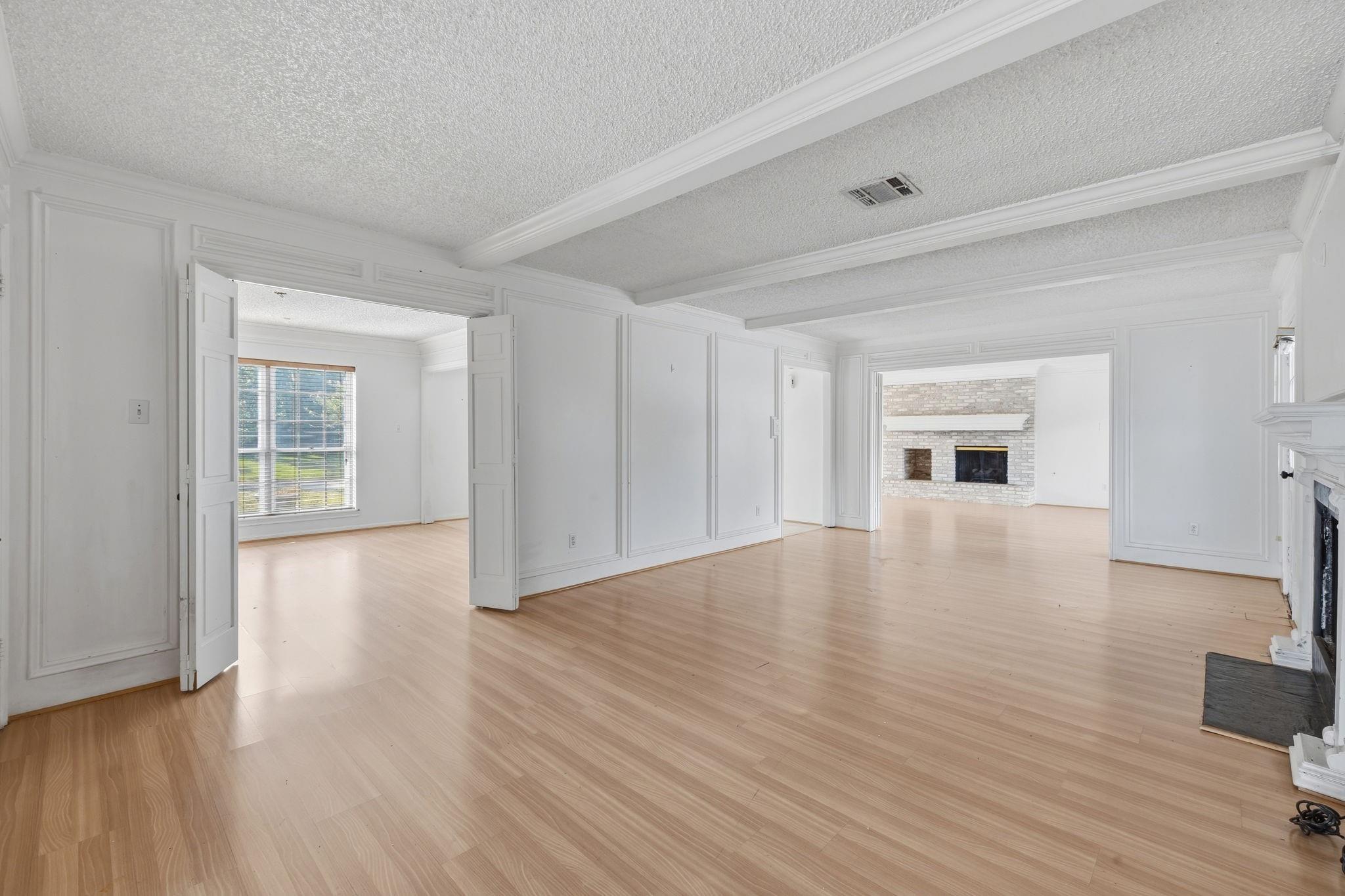 Vacant living room with pale wood floors, white paneled walls, and an adjoining room with a stone fireplace.