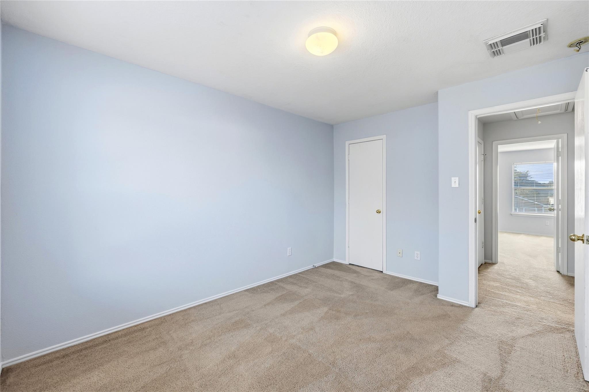 Empty bedroom with pale blue walls and beige carpet, viewed toward a doorway and adjoining room.