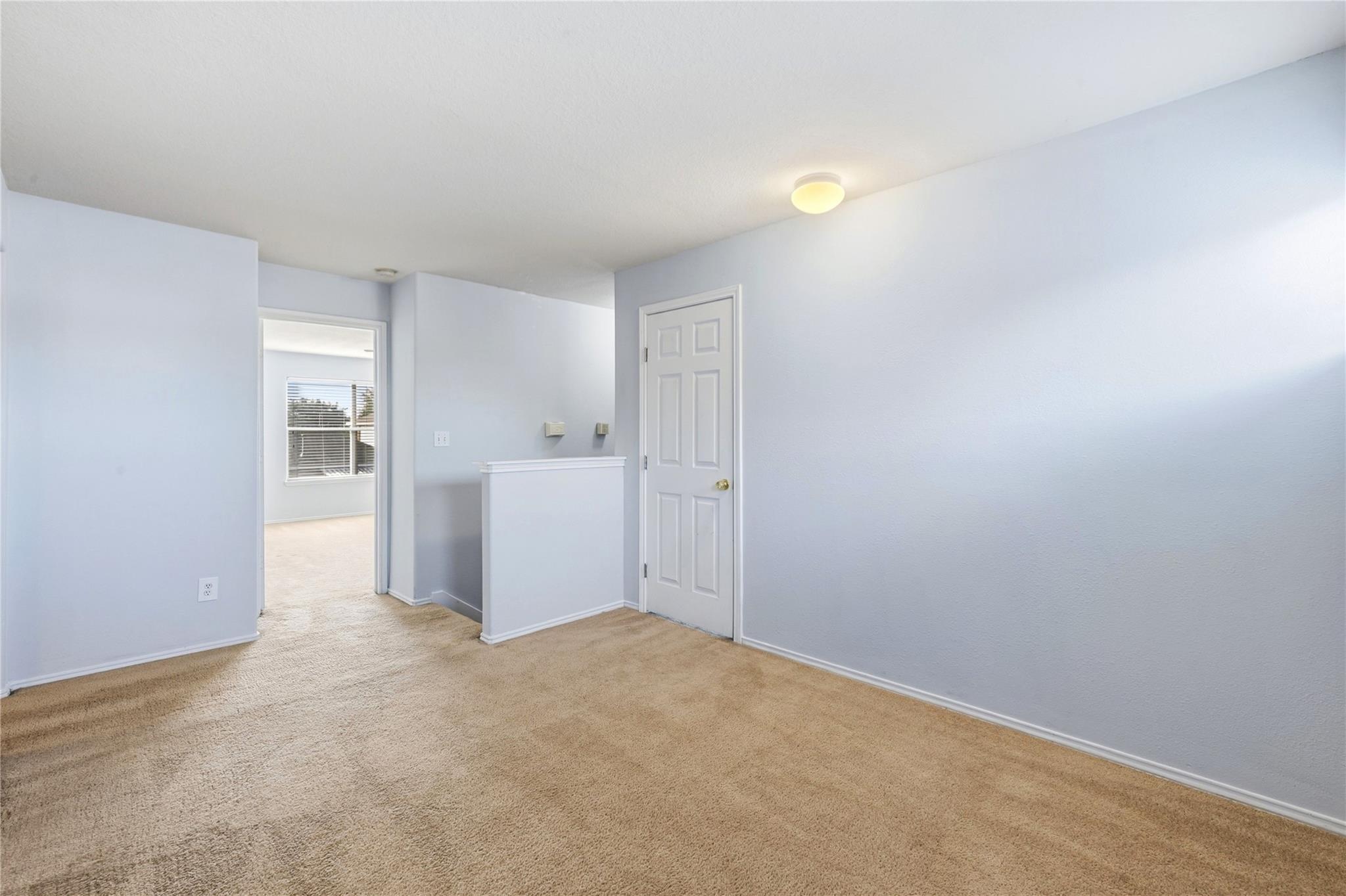 Empty carpeted room with pale walls, a white paneled door, and an adjoining room visible through the doorway.