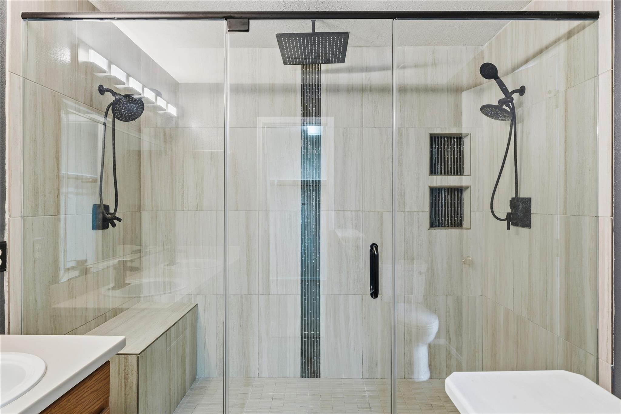 Bathroom with a glass-enclosed shower, dual black showerheads, tiled niches and a built-in bench.