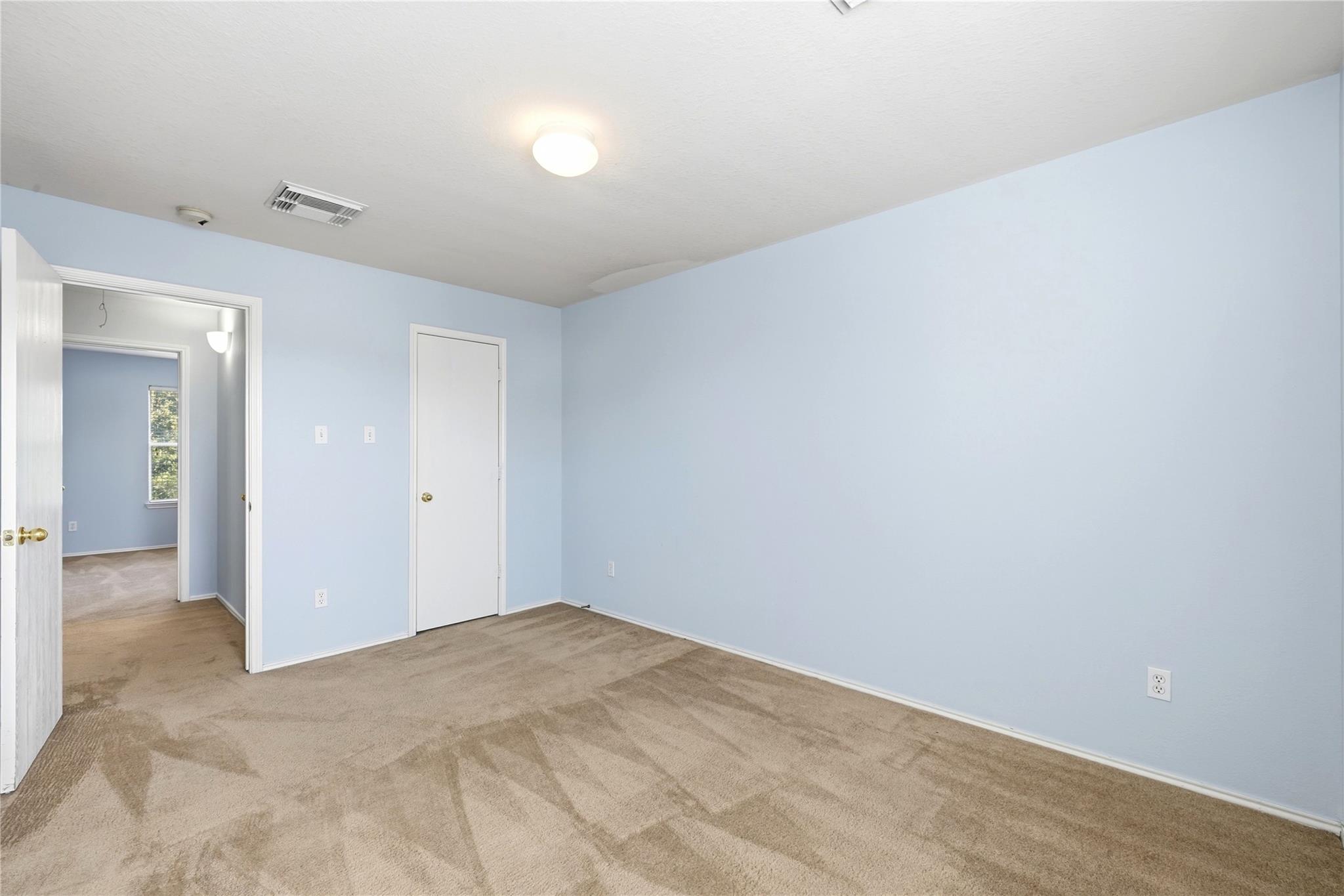 Empty bedroom with pale blue walls, beige carpet, and an open doorway to an adjoining room.